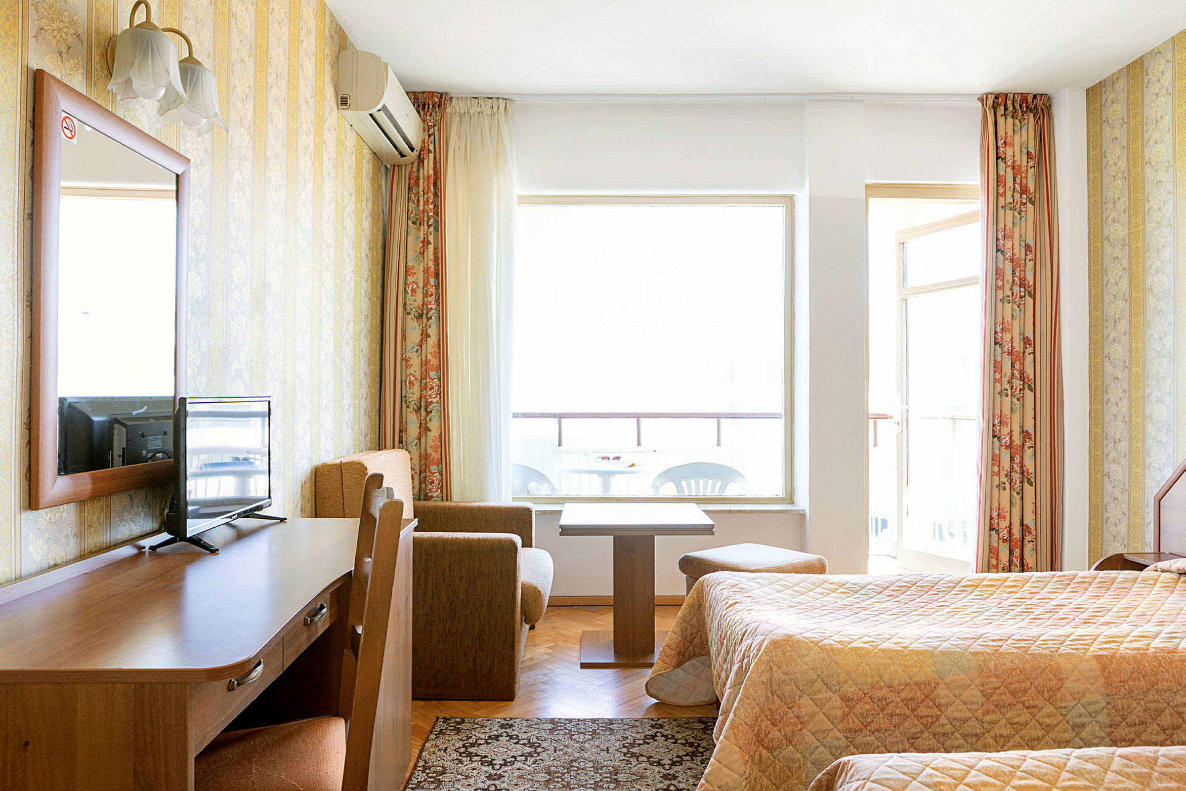 Ett hotellrum med två sängar, ett träskrivbord med en TV på, ett litet soffhörn med soffbord och en balkong i bakgrunden. (AI-genererad text)