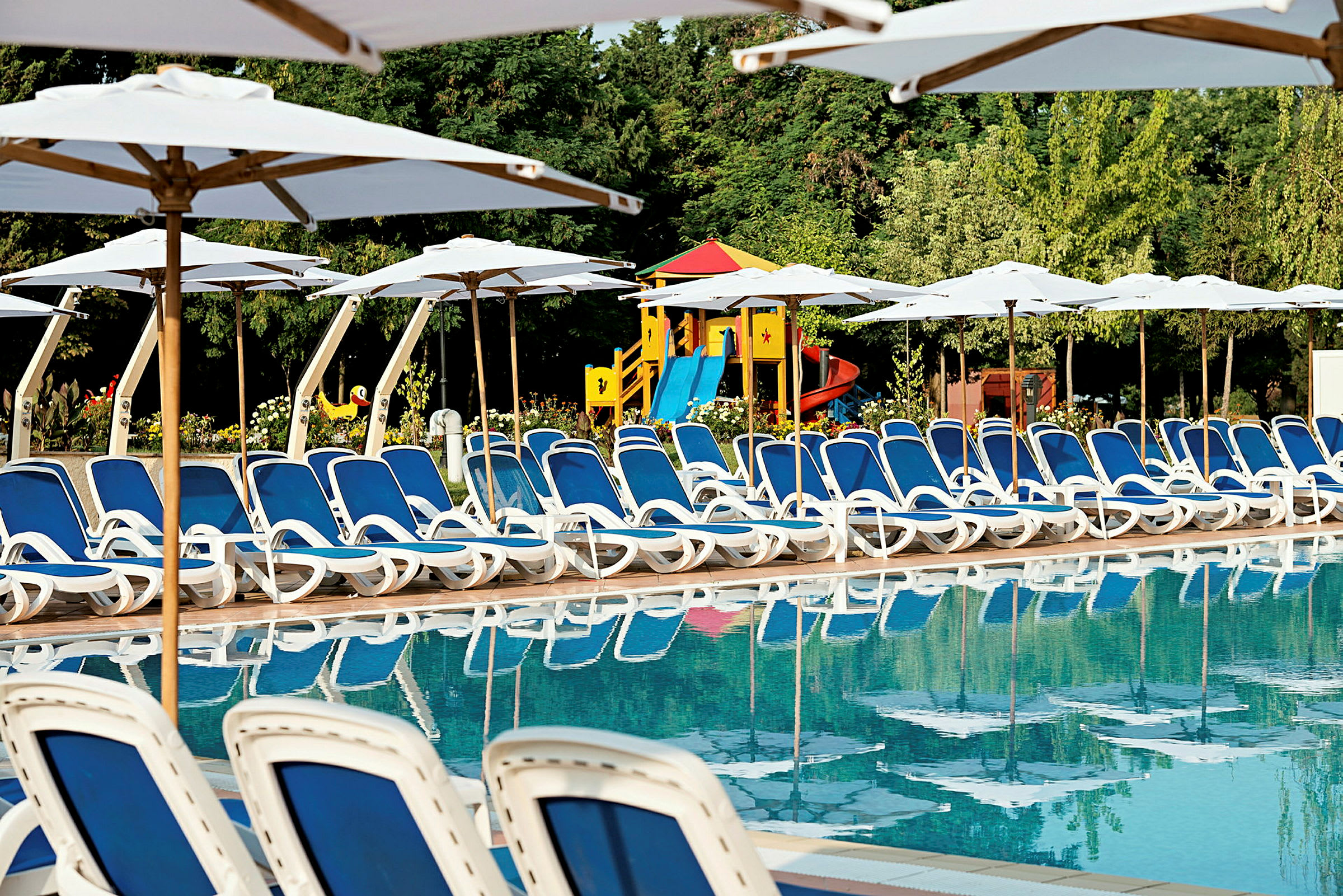 Bild av solstolar med blå dynor vid en swimmingpool. Bakom solstolarna syns vita parasoll och en lekplats för barn. (AI-genererad text)