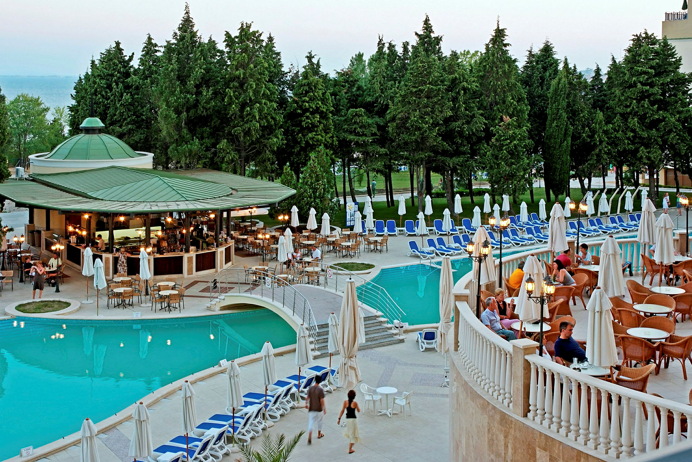Bild av ett hotellområde med en pool, omgivande solstolar och ett utomhuscafé med sittplatser. Tallar och grönska i bakgrunden. (AI-genererad text)