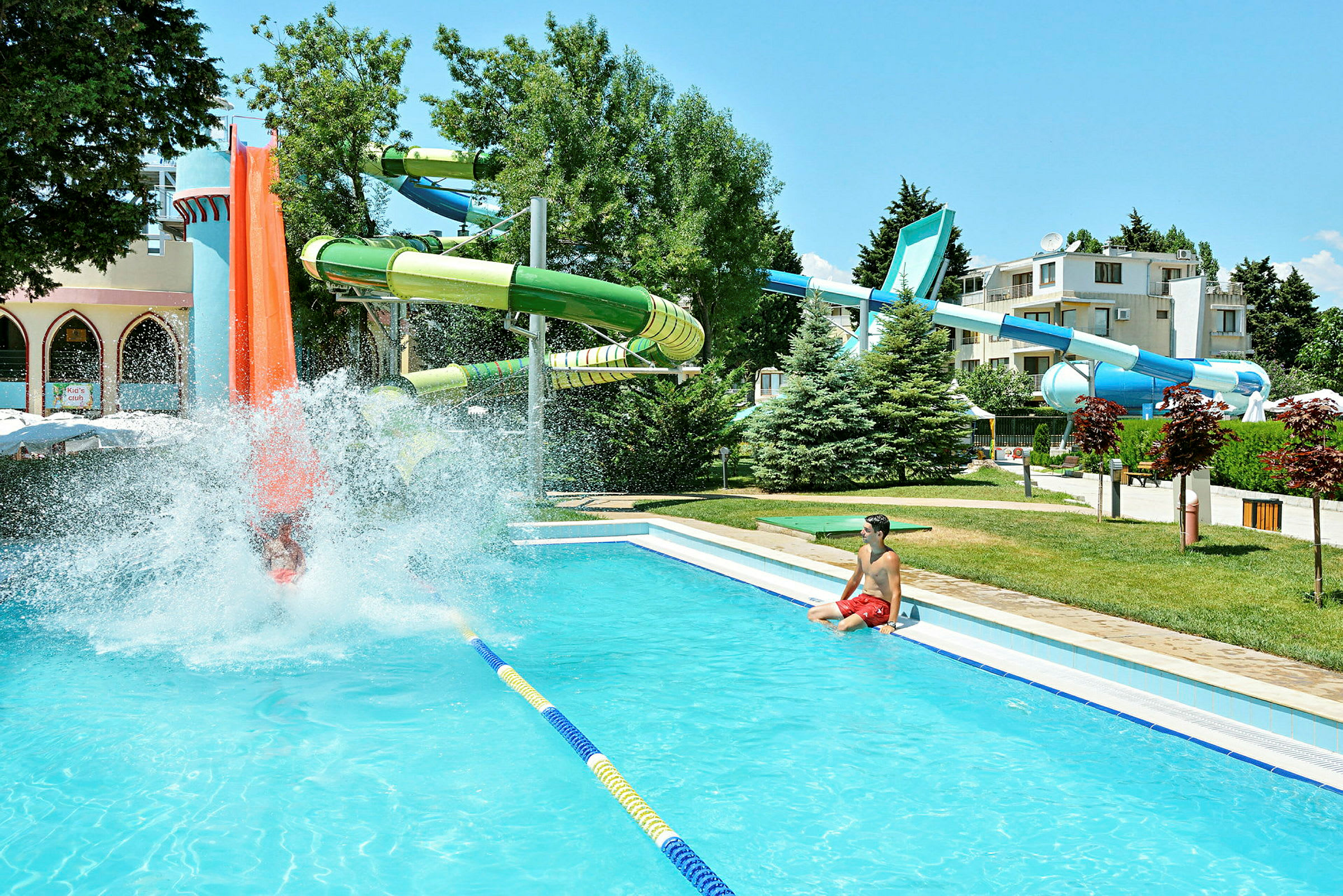 En vattenpark med rutschkanor och en pool, omgiven av grönska och byggnader. (AI-genererad text)
