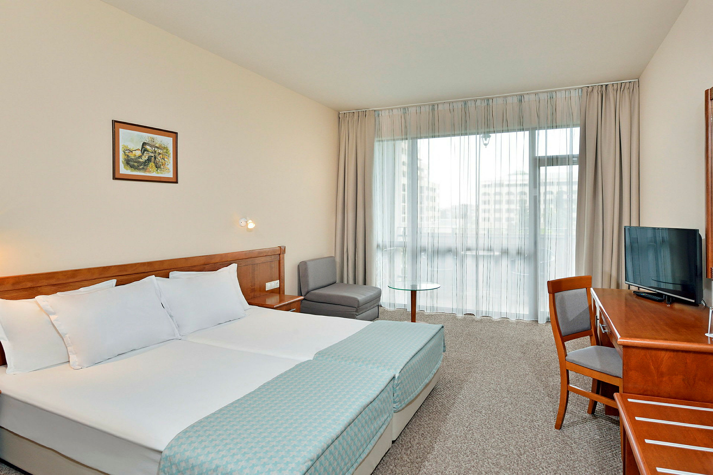 Ett hotellrum med en dubbelsäng, skrivbord, stol, TV och sittgrupp. Ett stort fönster släpper in dagsljus. (AI-genererad text)