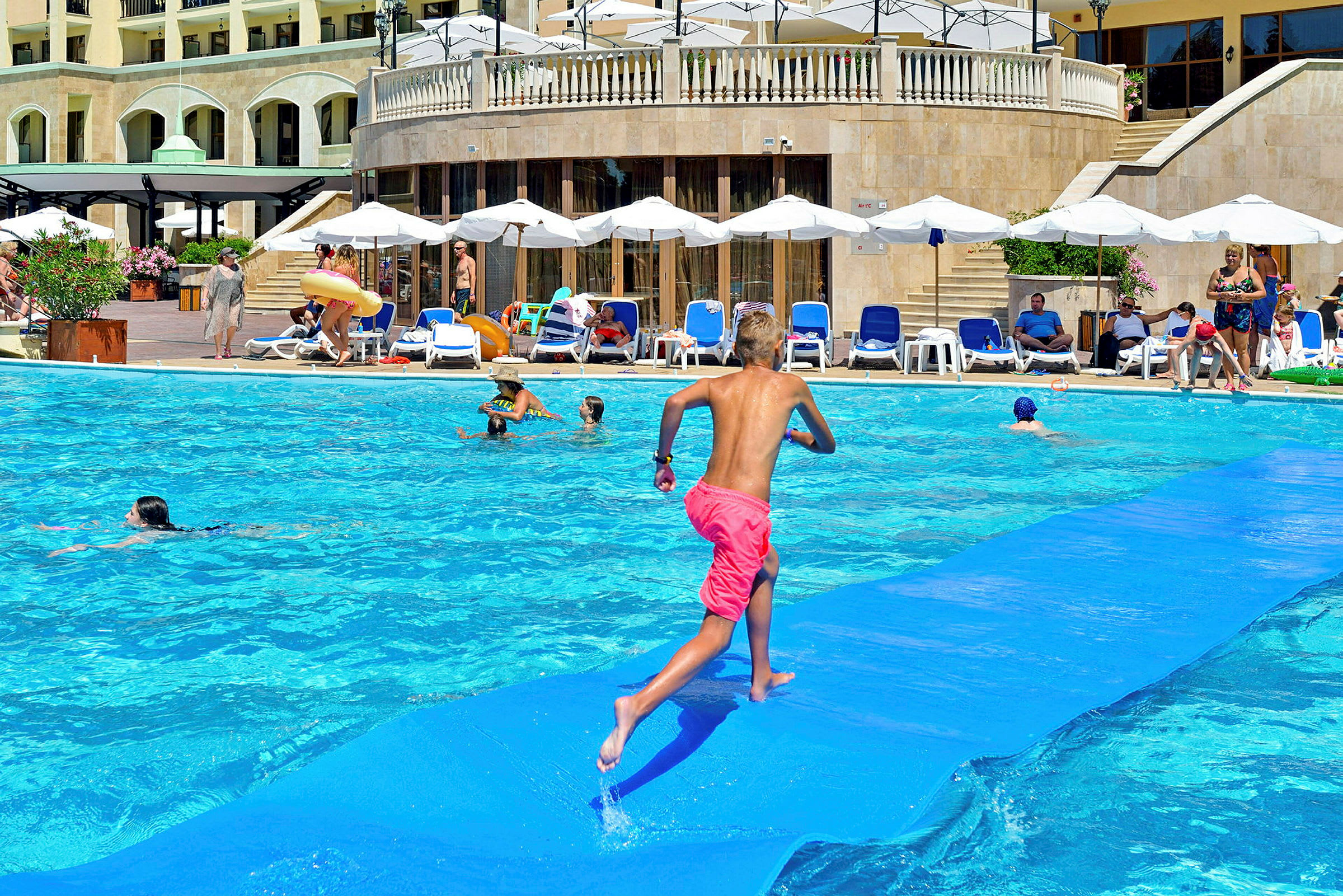 Barn springer på matta som flyter i en swimmingpool medan vuxna och barn kopplar av vid poolkanten. (AI-genererad text)