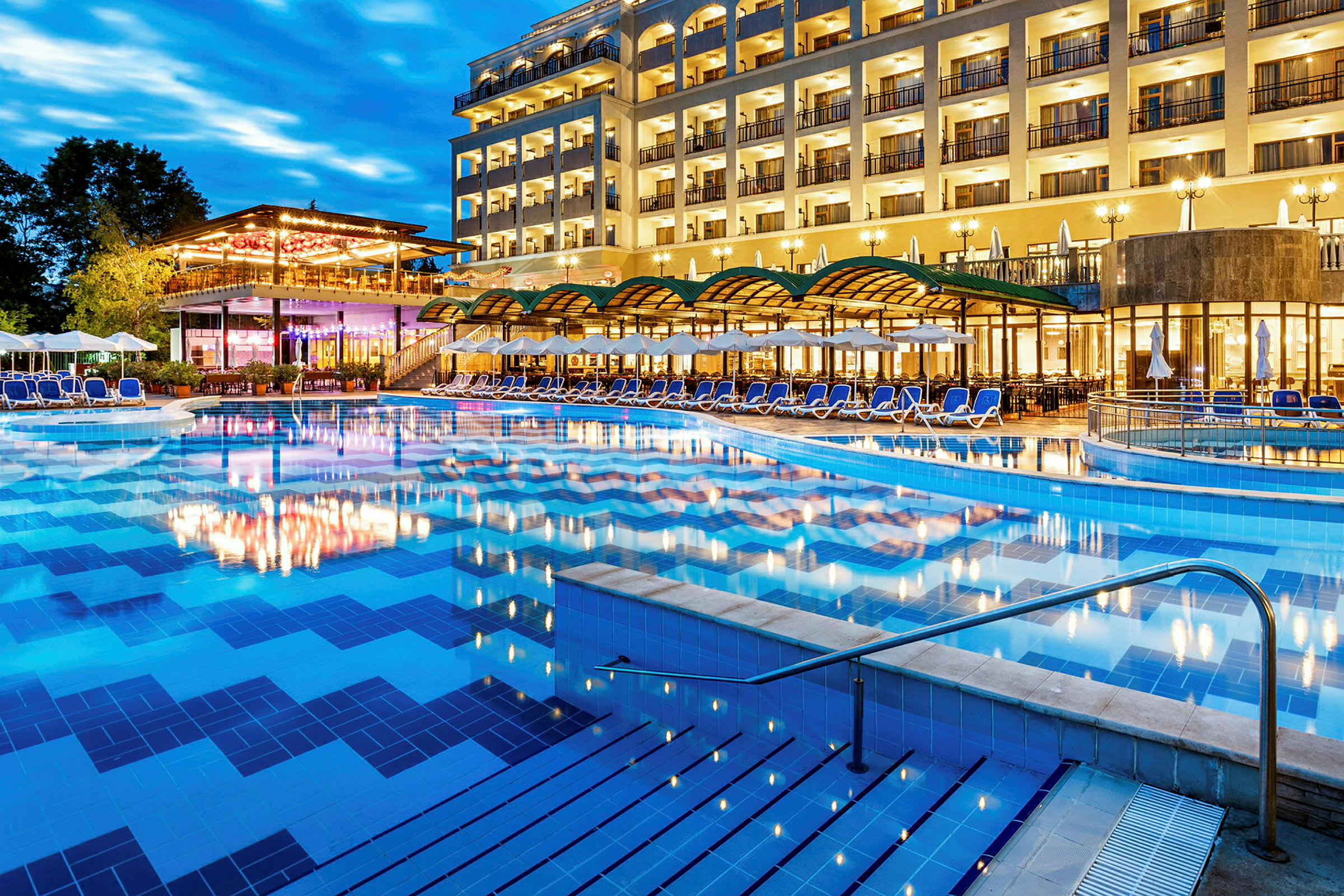 Ett hotell med många balkonger och områden för avslappning vid en stor belyst utomhuspool. (AI-genererad text)