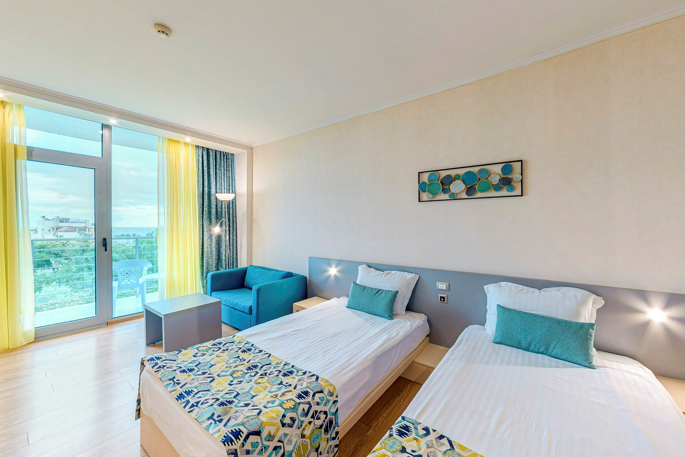 Ett hotellrum med två enkelsängar, en blå fåtölj, och ett bord. Rummet har ljusa väggar och ett stort fönster med gul gardin och balkong. (AI-genererad text)