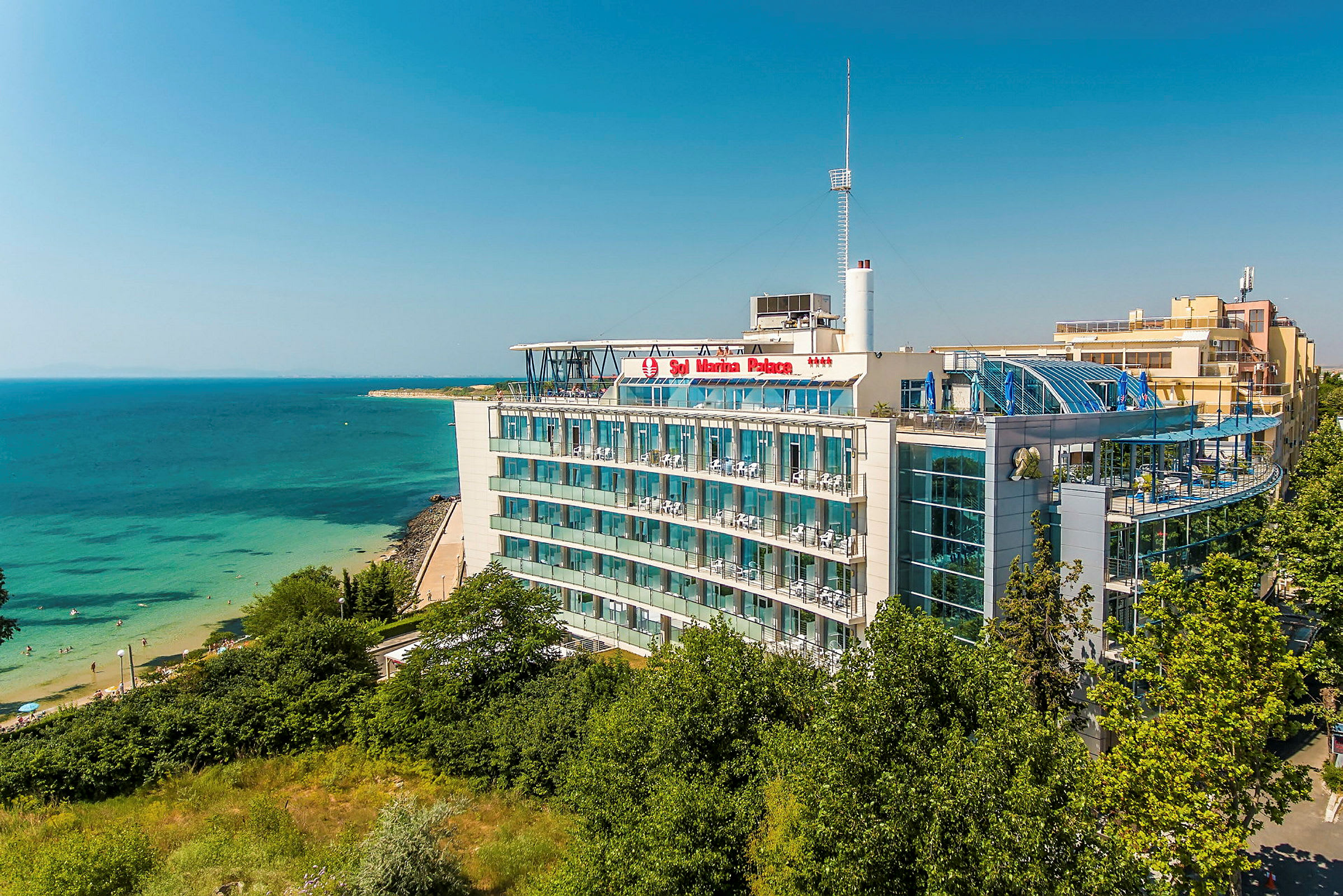Ett hotell vid havet med balkonger och glasfasader, omgiven av grönska, med en strand i närheten. (AI-genererad text)
