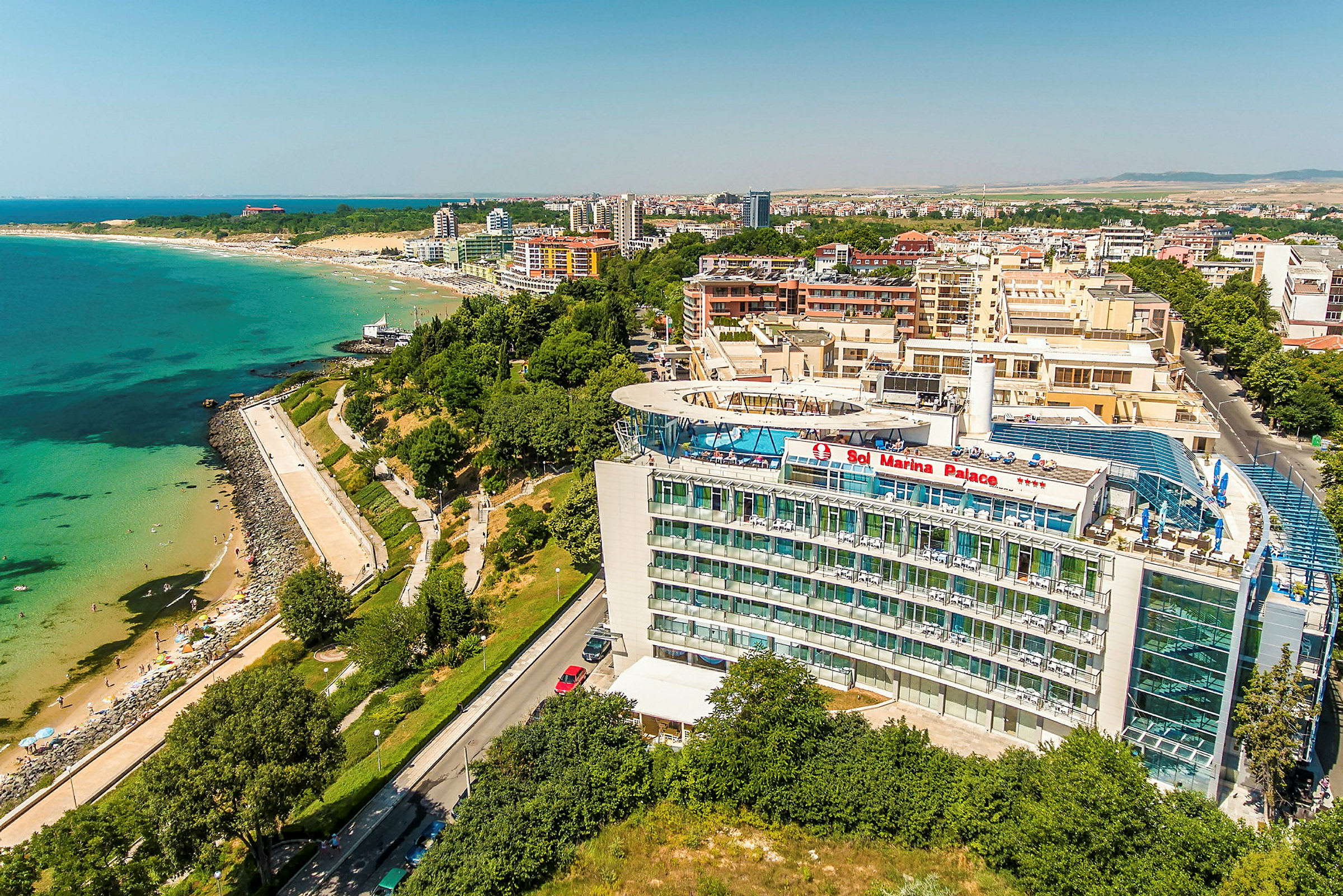 Flygfoto av ett hotell med texten ”Sol Marina Palace”, belägen nära en strand och kustlinje med grönska och byggnader i bakgrunden. (AI-genererad text)