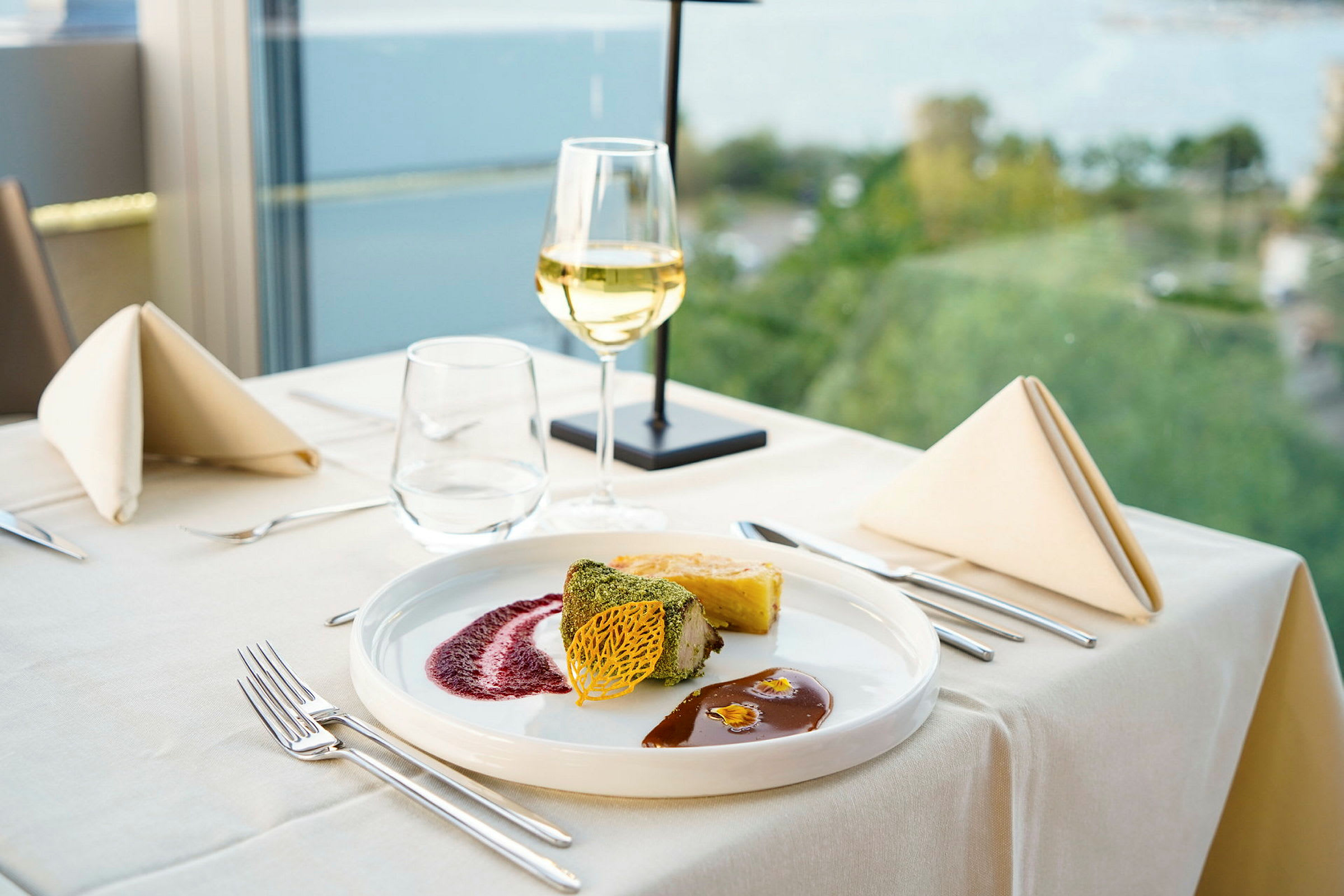 En elegant dukad tallrik med en maträtt som innehåller en grön örtkrödd bit, en potatisbakelse och såser. Ett glas vitt vin står på bordet med en bakgrund av fönster med utsikt mot natur. (AI-genererad text)