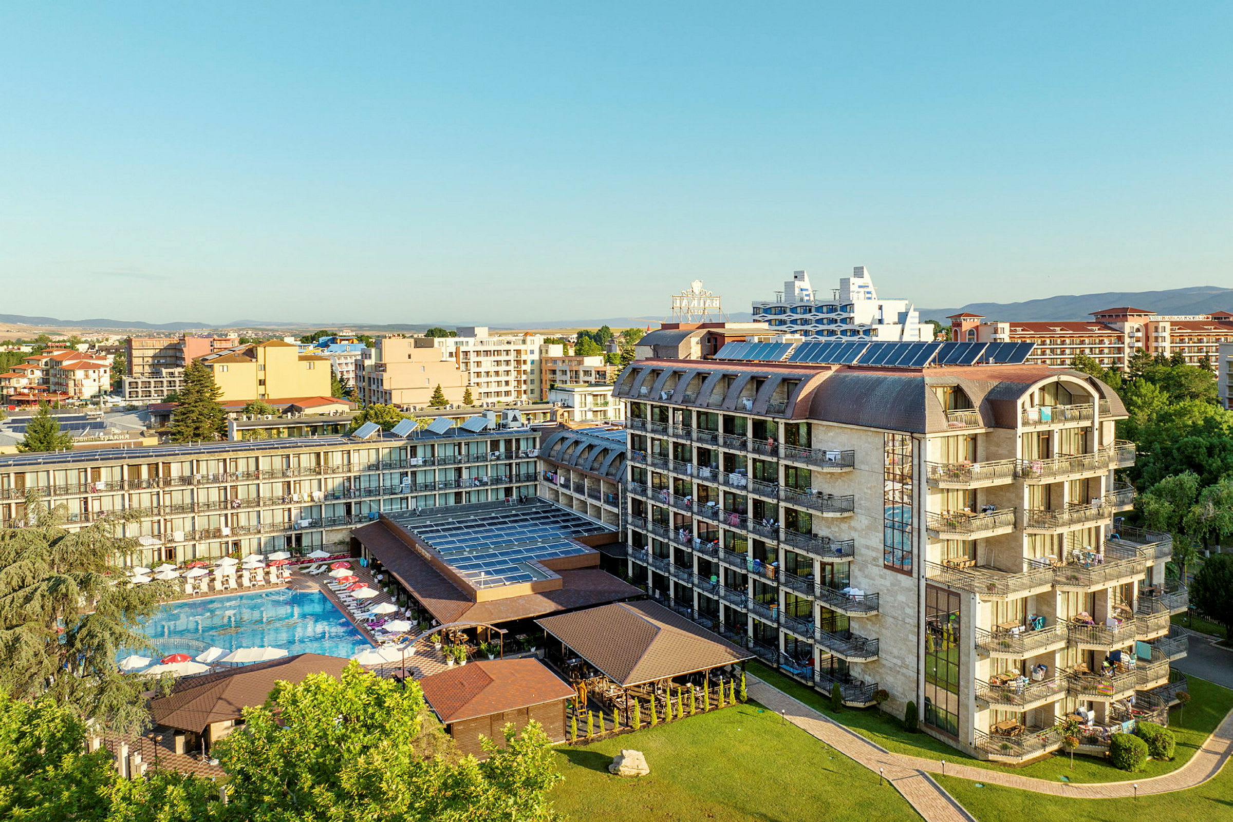 Flygfoto av ett hotell med pool och solstolar, omgiven av andra byggnader och grönska. (AI-genererad text)