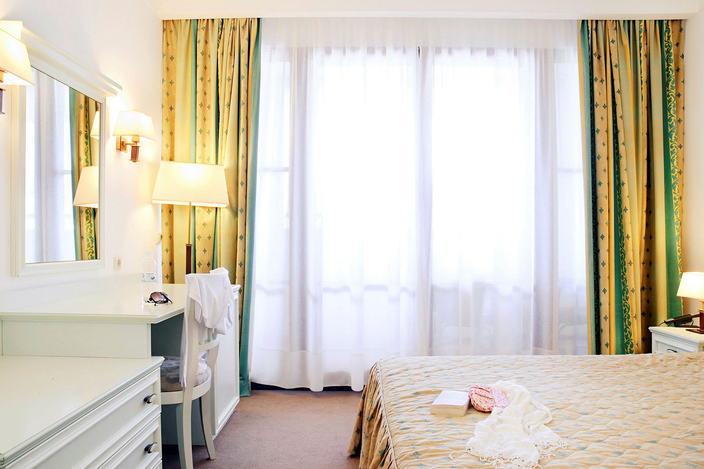Ett ljust hotellrum med en säng, en spegel och gardiner som täcker ett stort fönster. (AI-genererad text)
