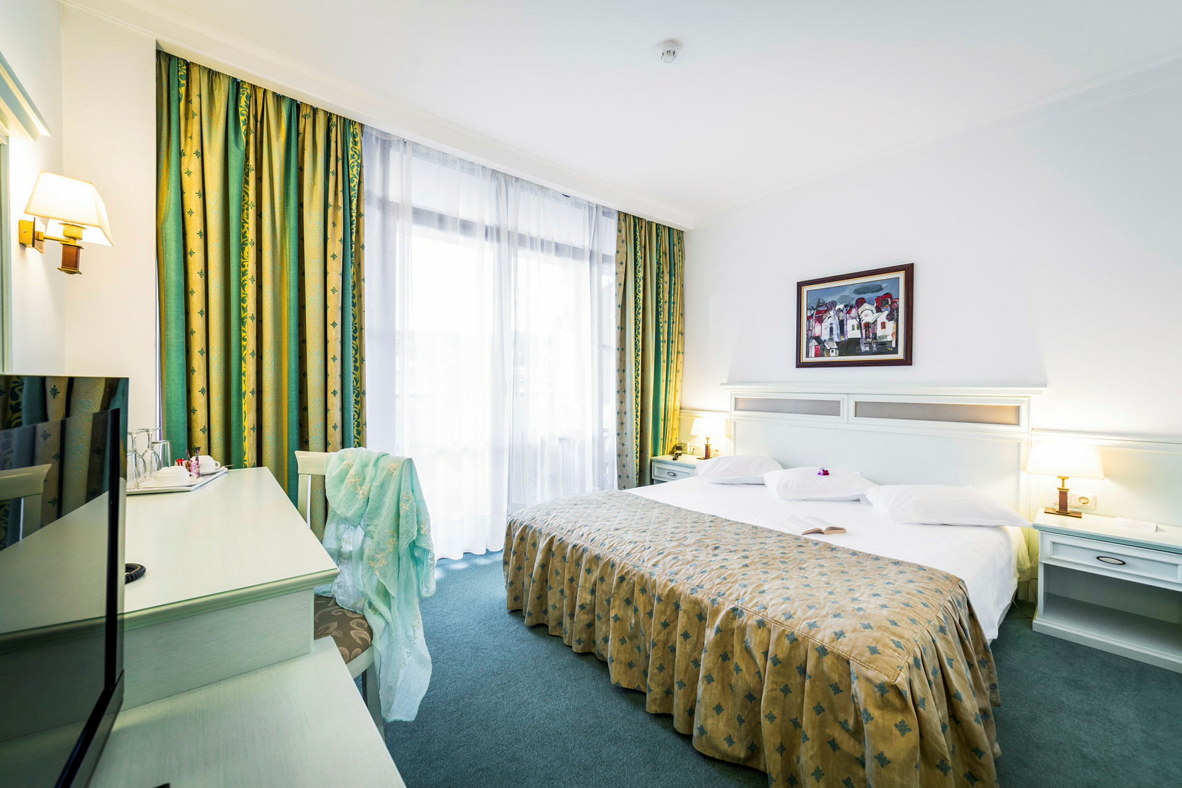 Ett hotellrum med en stor säng, vita sängkläder och ett mönstrat överkast. Det finns bordslampor bredvid sängen och ett skrivbord med en stol i hörnet. (AI-genererad text)