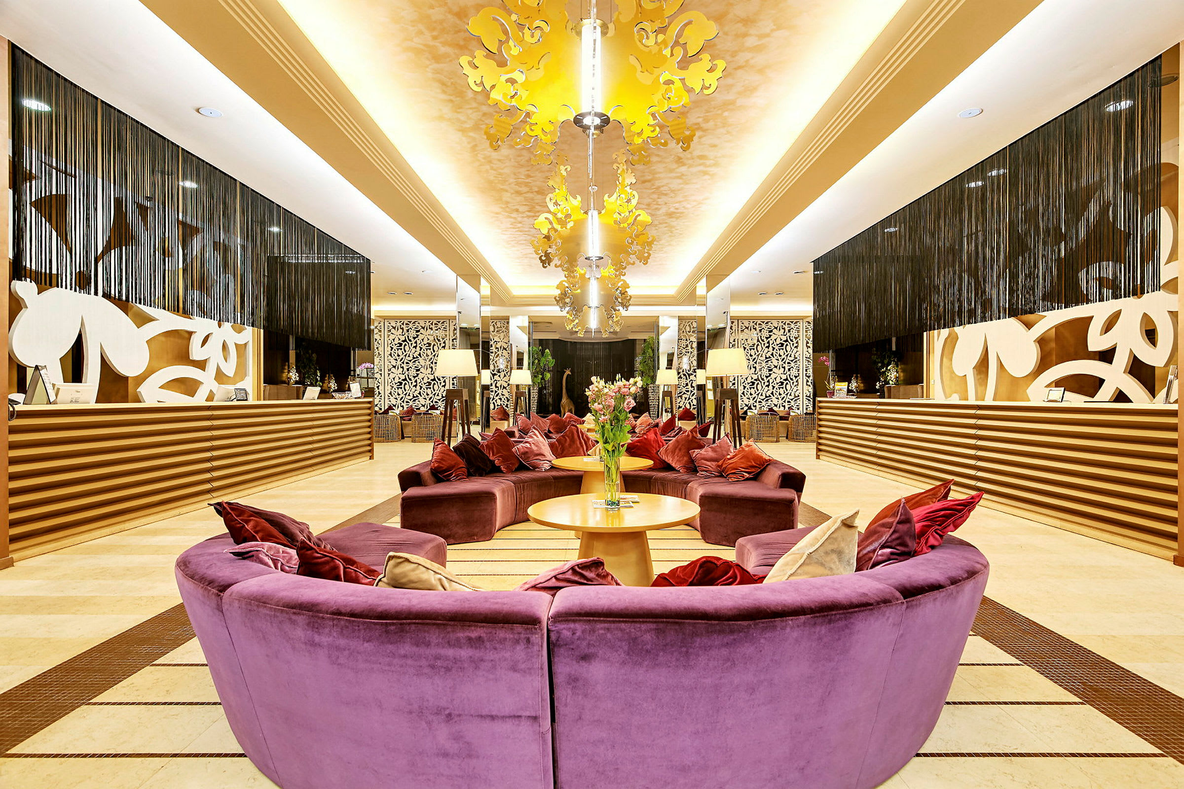 Receptionen på ett hotell med en elegant inredning. Färggranna soffor i lila och kuddar är centrerade med två bord och blommor i mitten. Bakom receptionen syns dekorativa mönster och hängande gardiner. (AI-genererad text)