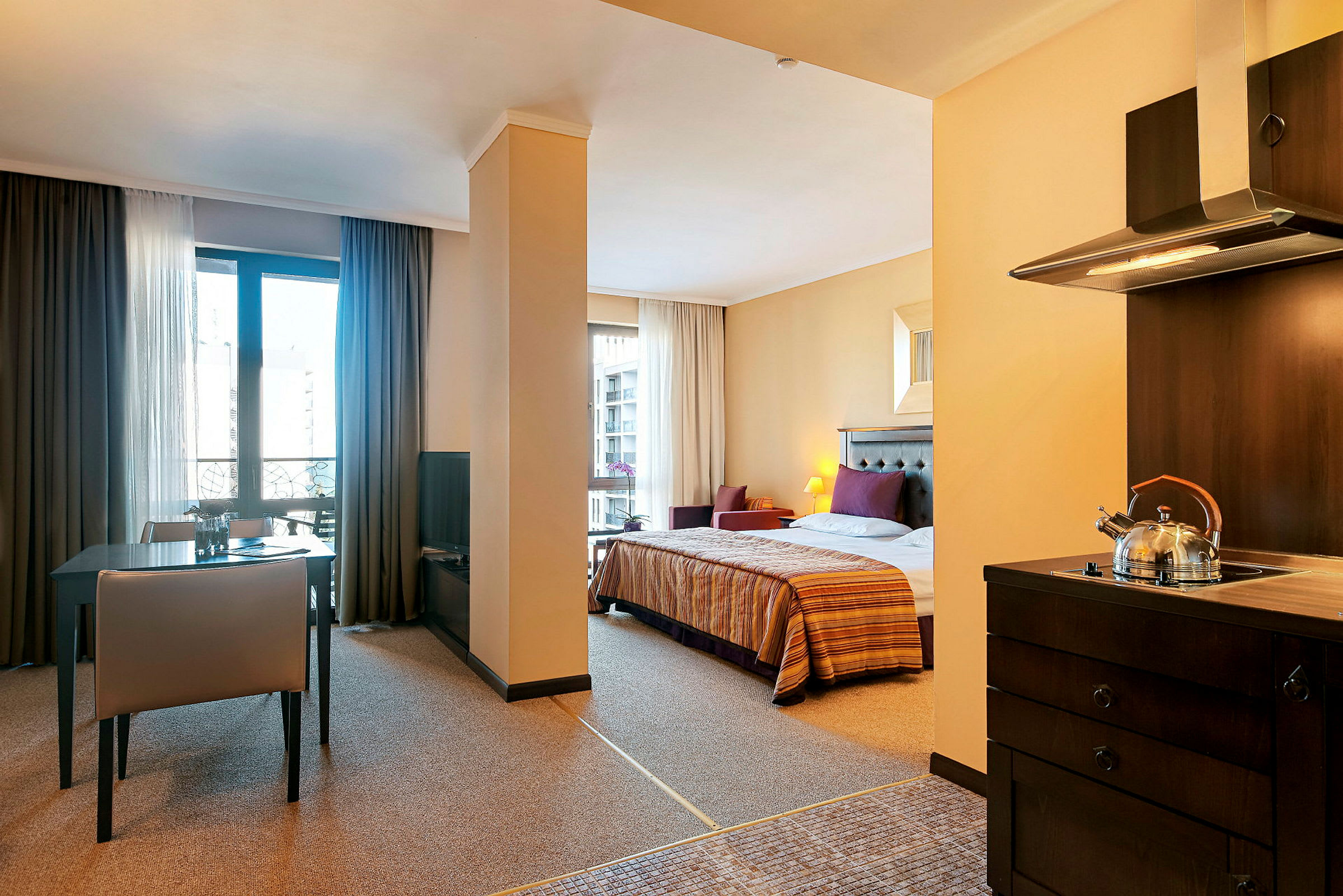 Interiör av ett hotellrum med sovplats, matbord och kök. Sängen är bäddad och köket har en tekanna på spisen. (AI-genererad text)