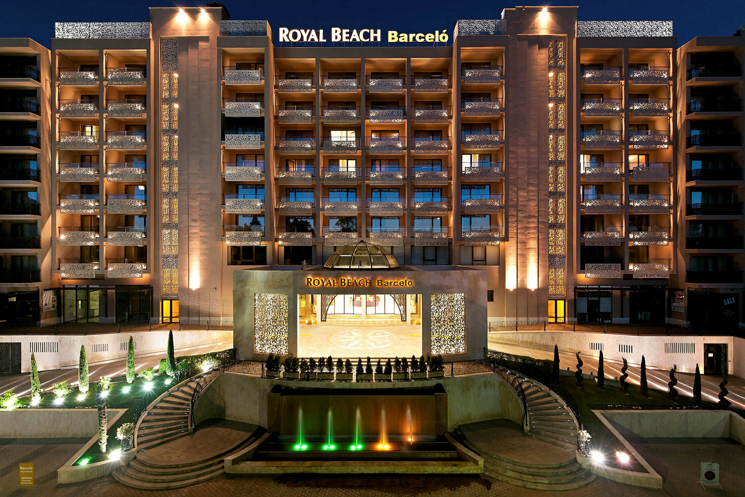 En nattbild av ett upplyst hotell med namnet Royal Beach Barceló, med en fontän och trappor i förgrunden. (AI-genererad text)