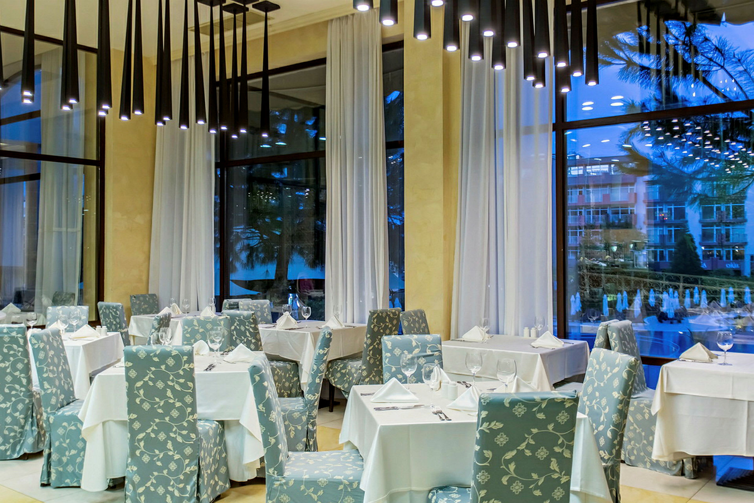 En elegant restaurang med bord klädda i vita dukar och stolar med blommigt mönster, höga fönster med vita gardiner. (AI-genererad text)