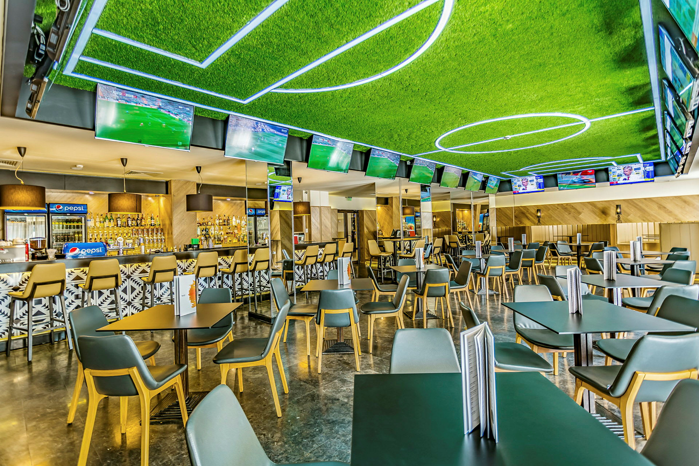 Ett rum med flera bord och stolar, en bar i bakgrunden och tv-skärmar som visar fotbollsmatcher. (AI-genererad text)