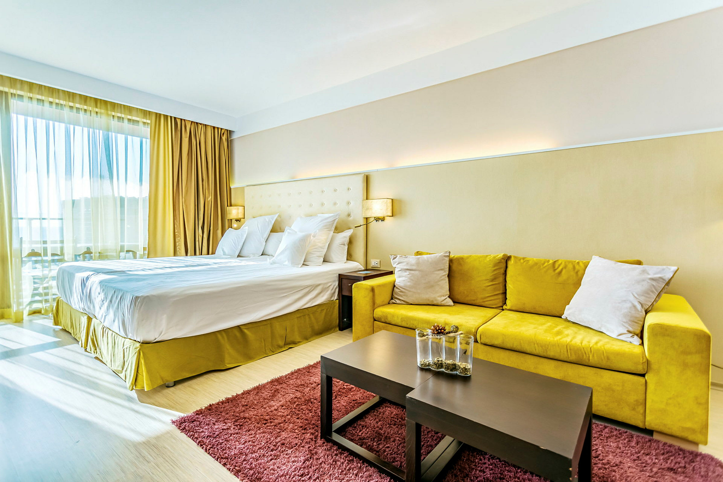 Ett hotellrum med en stor dubbelsäng, gul soffa, ett bord med dekorativ vas och en röd matta. (AI-genererad text)