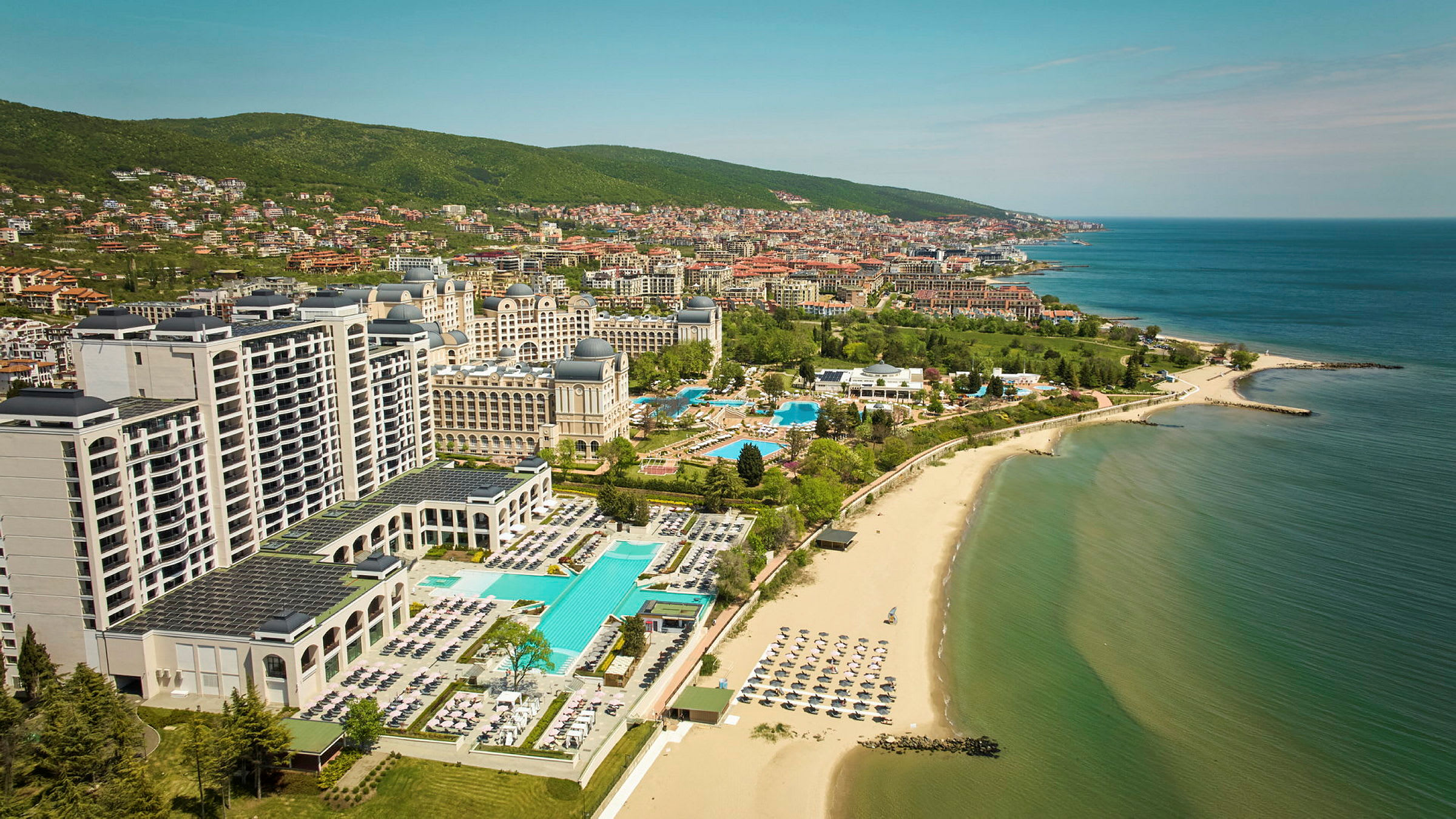 Hotell och pooler vid stranden med havet och staden i bakgrunden. (AI-genererad text)