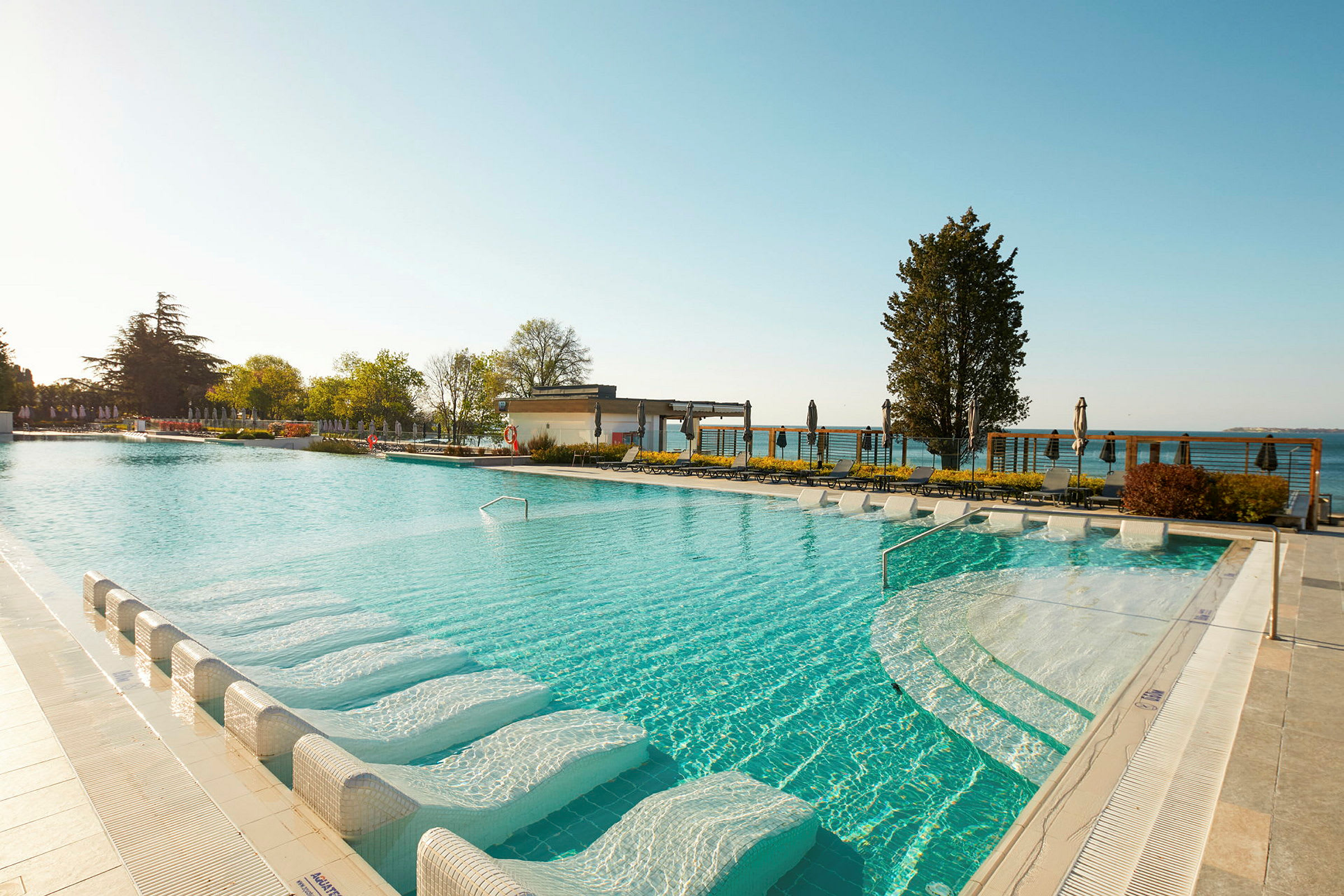 Modern pool med liggstolar i vattnet och utsikt över omgivande grönska och himmel. (AI-genererad text)