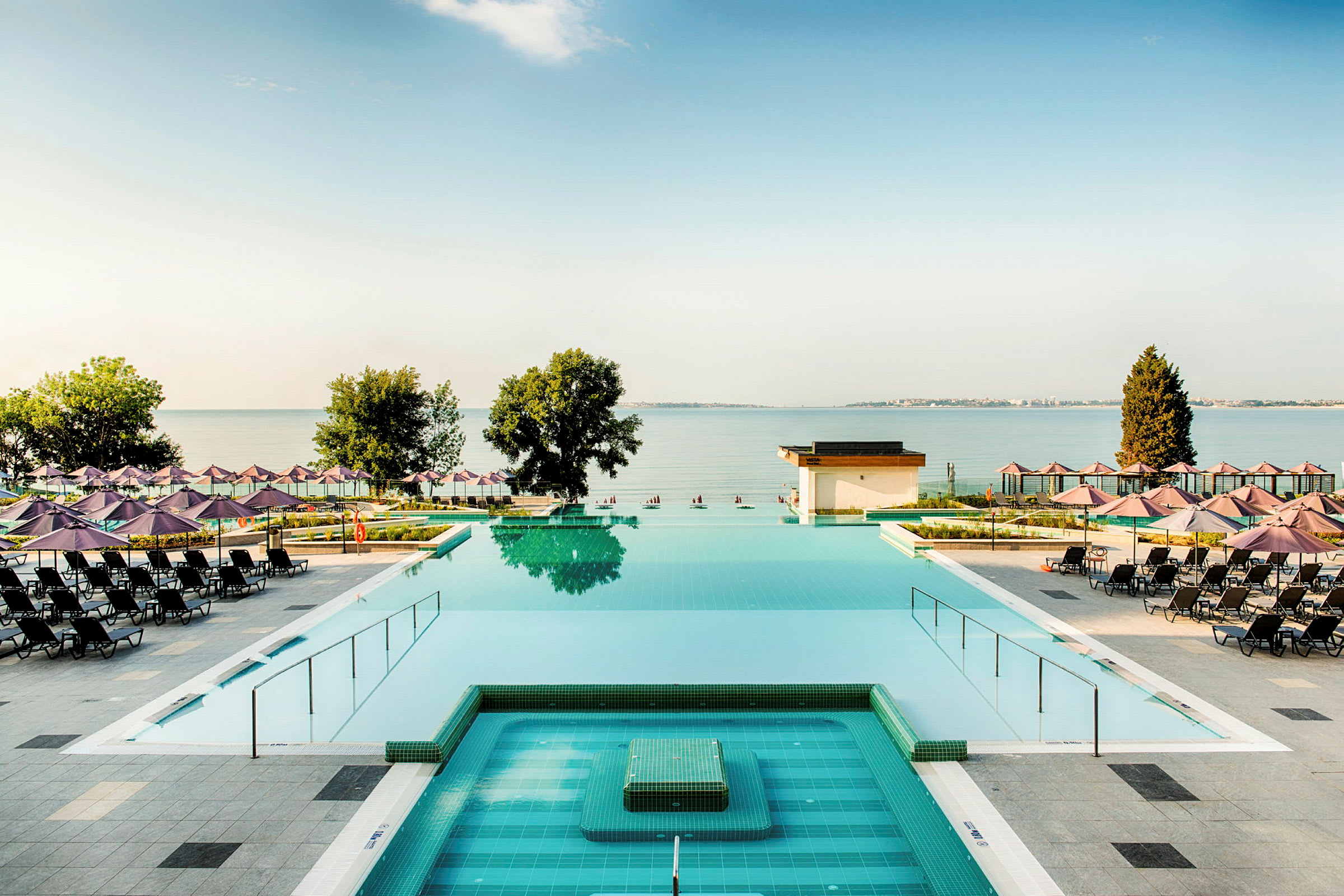 En utsikt över en pool med solstolar och parasoller utspridda runt omkring, samt havet och träd i bakgrunden. (AI-genererad text)
