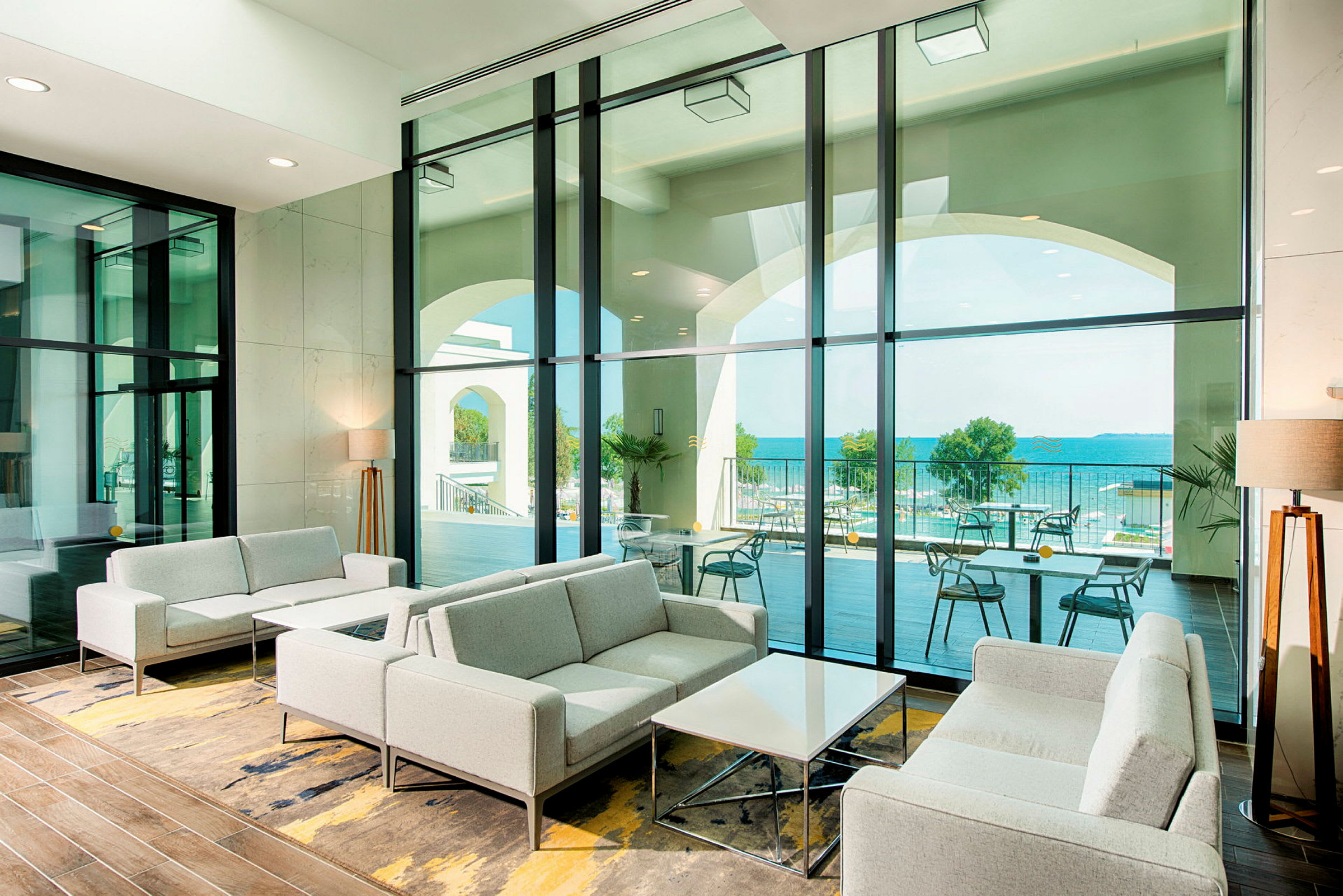 En modern lounge med grå soffor och stora fönster som visar utsikt över en terrass och havet. (AI-genererad text)