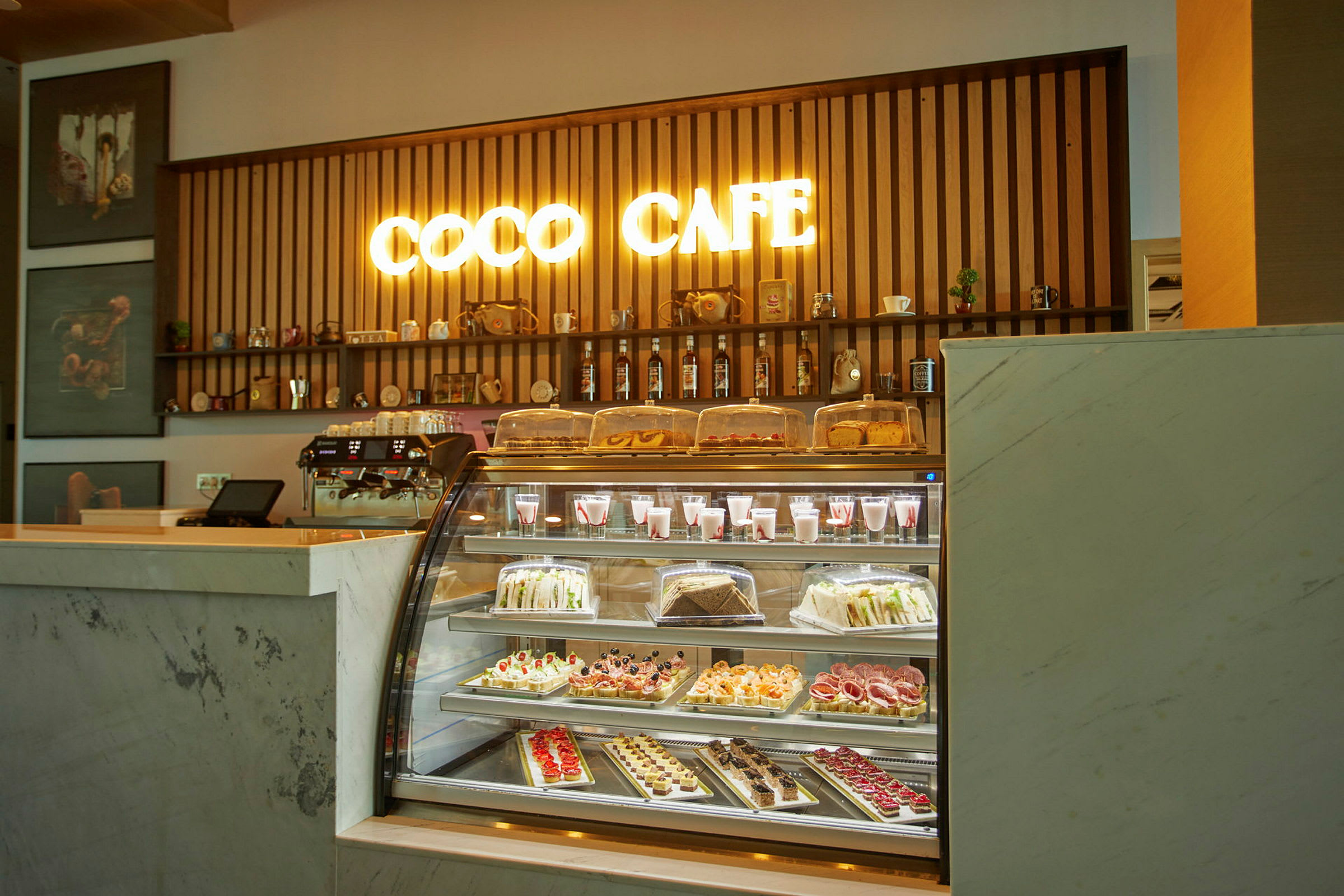 En kafédisk med en belyst skylt som säger 'Coco Cafe' och ett urval av bakverk och drycker bakom glas. (AI-genererad text)