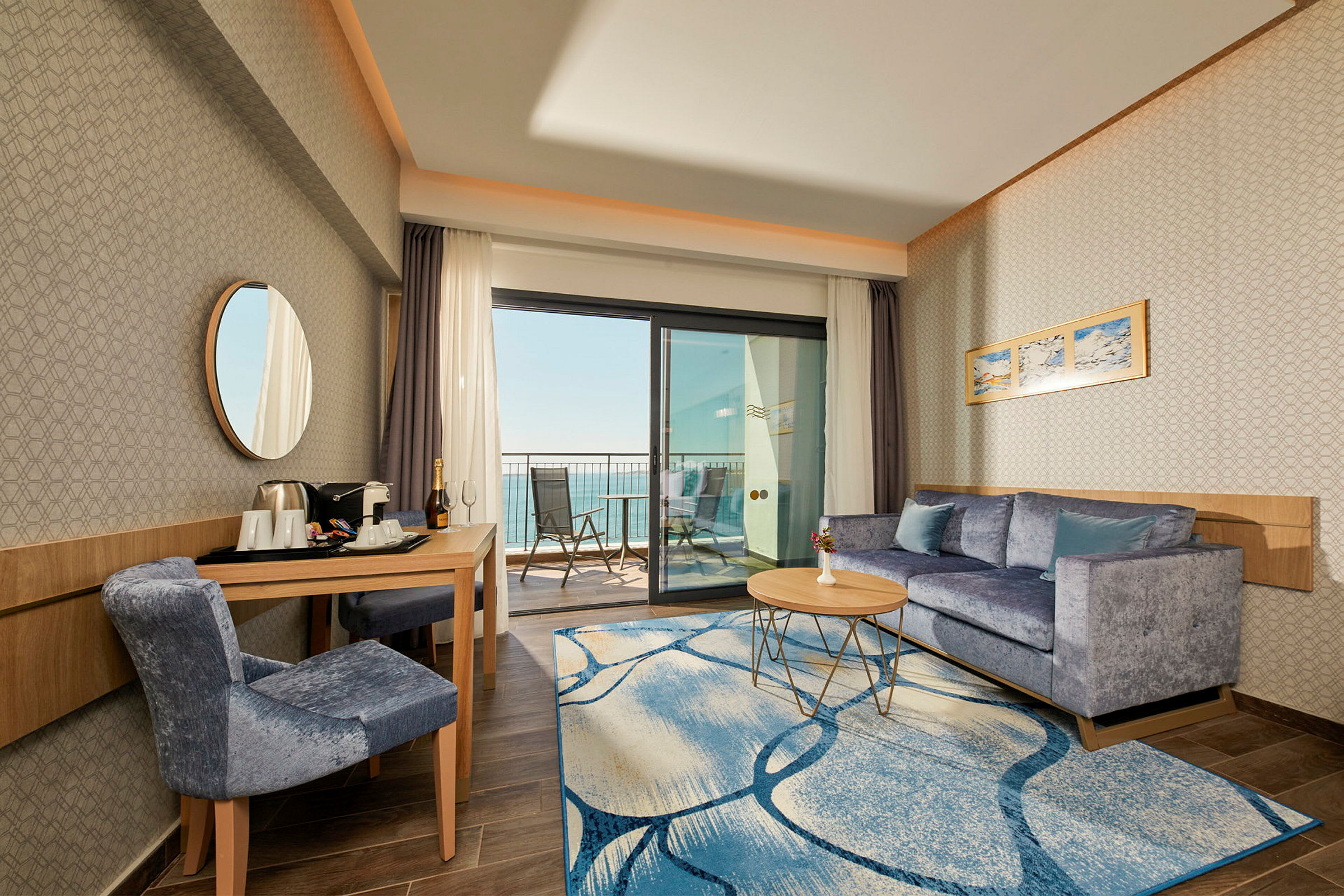 Hotellrum med sittgrupp, bord och stol samt balkong med utsikt mot havet. (AI-genererad text)