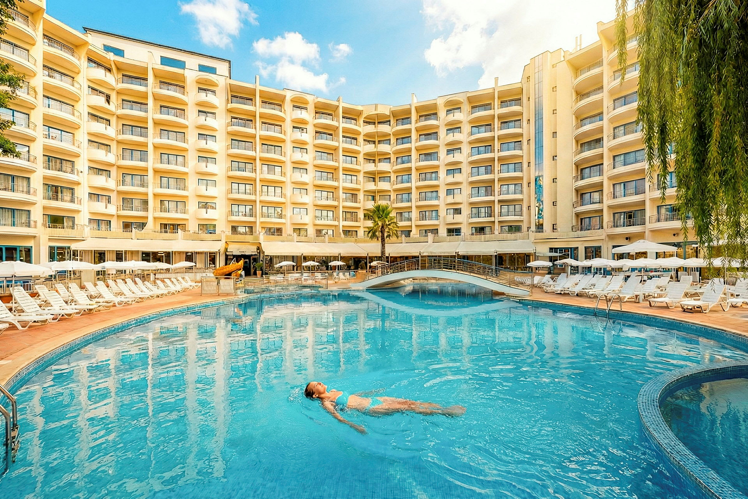 En stor utomhuspool med solstolar runt, framför ett hotell med flera våningar. En person simmar i poolen. (AI-genererad text)