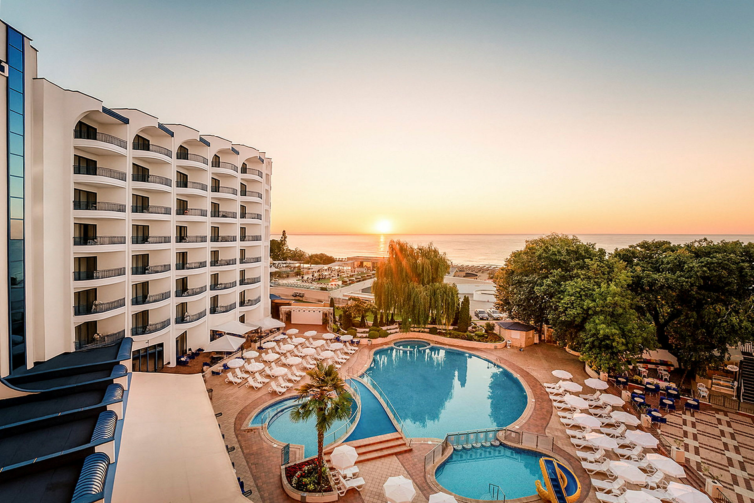 Bild av ett hotellområde vid solnedgången med en stor pool omgiven av parasoller och solstolar, samt havet i bakgrunden. (AI-genererad text)