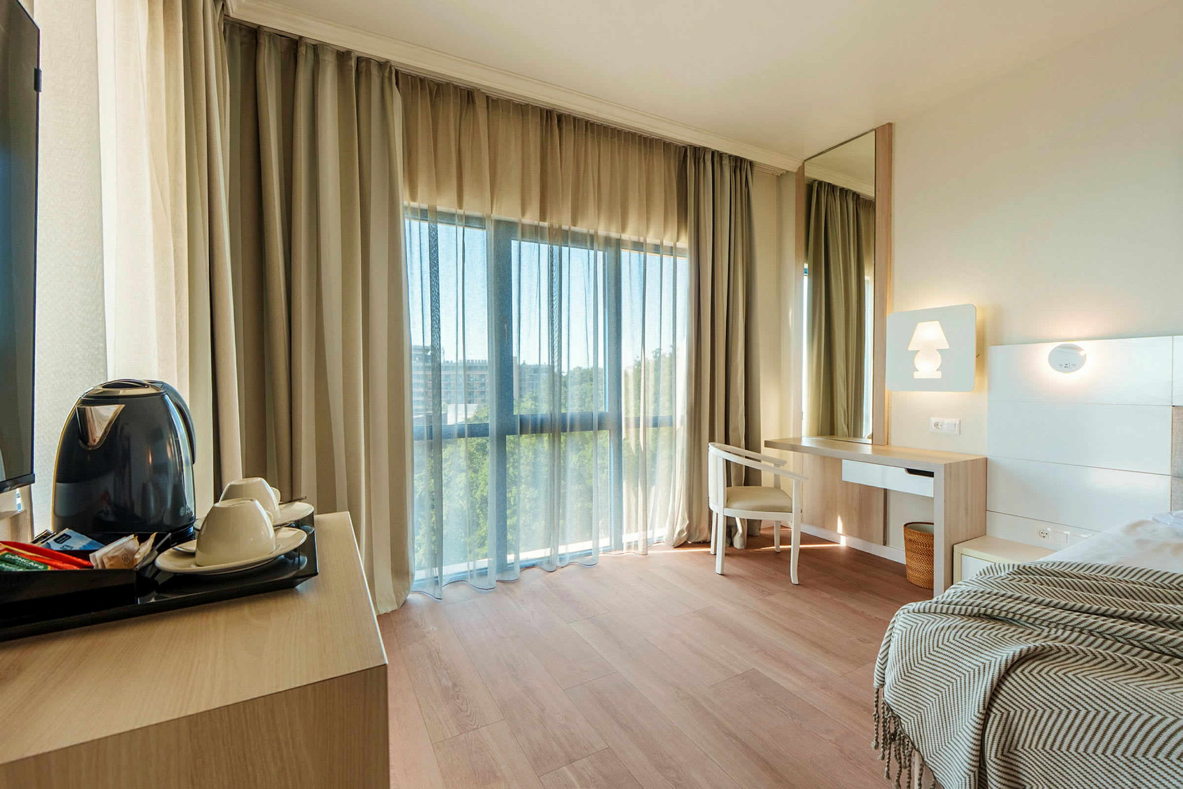 Ett ljust hotellrum med trägolv, ett skrivbord med stol och spegel, samt en säng med en filt. Ett stort fönster med gardiner släpper in dagsljuset. (AI-genererad text)