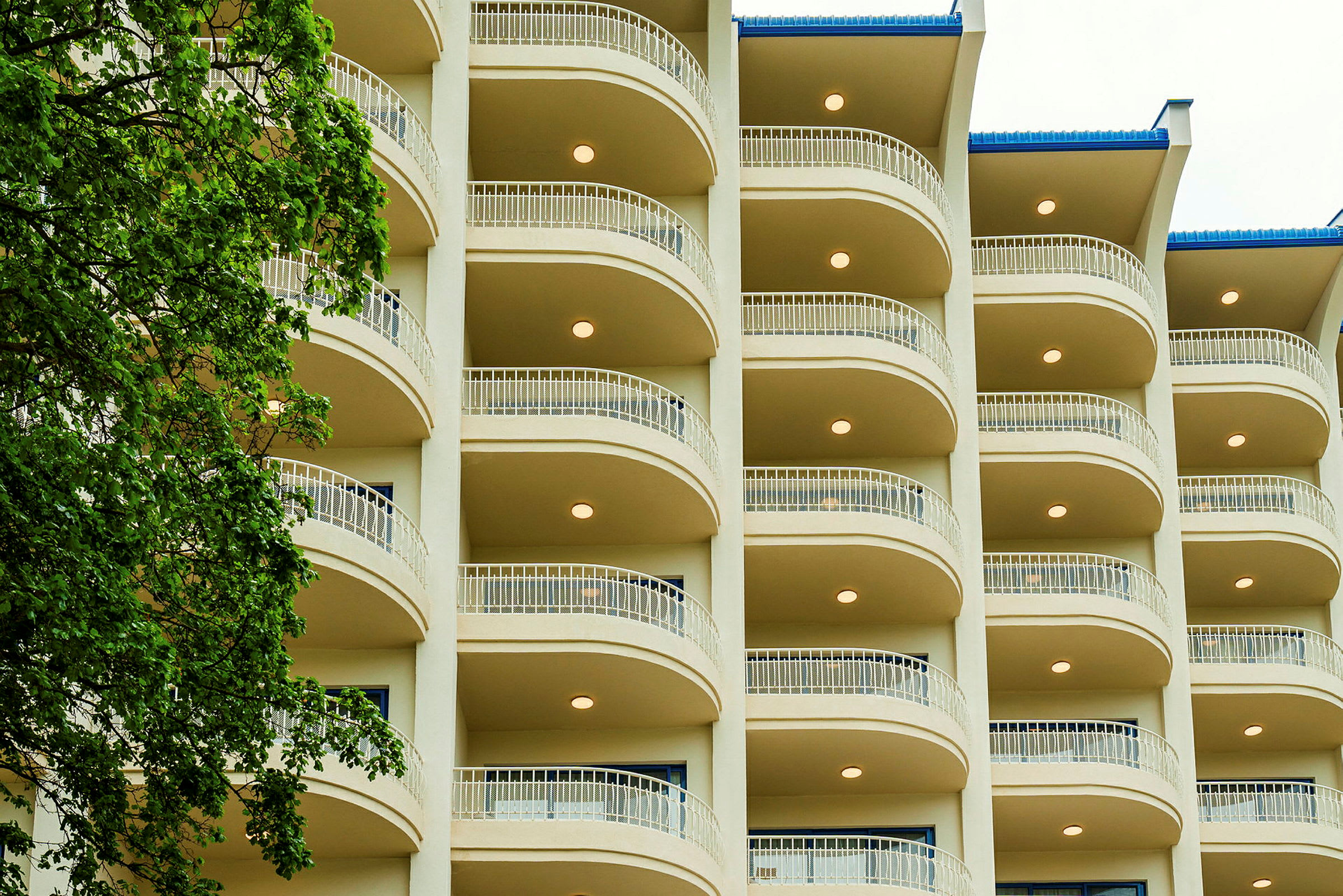 Visar fasaden på ett hotell med våningar och balkonger. Ett träd befinner sig till vänster i bilden. (AI-genererad text)