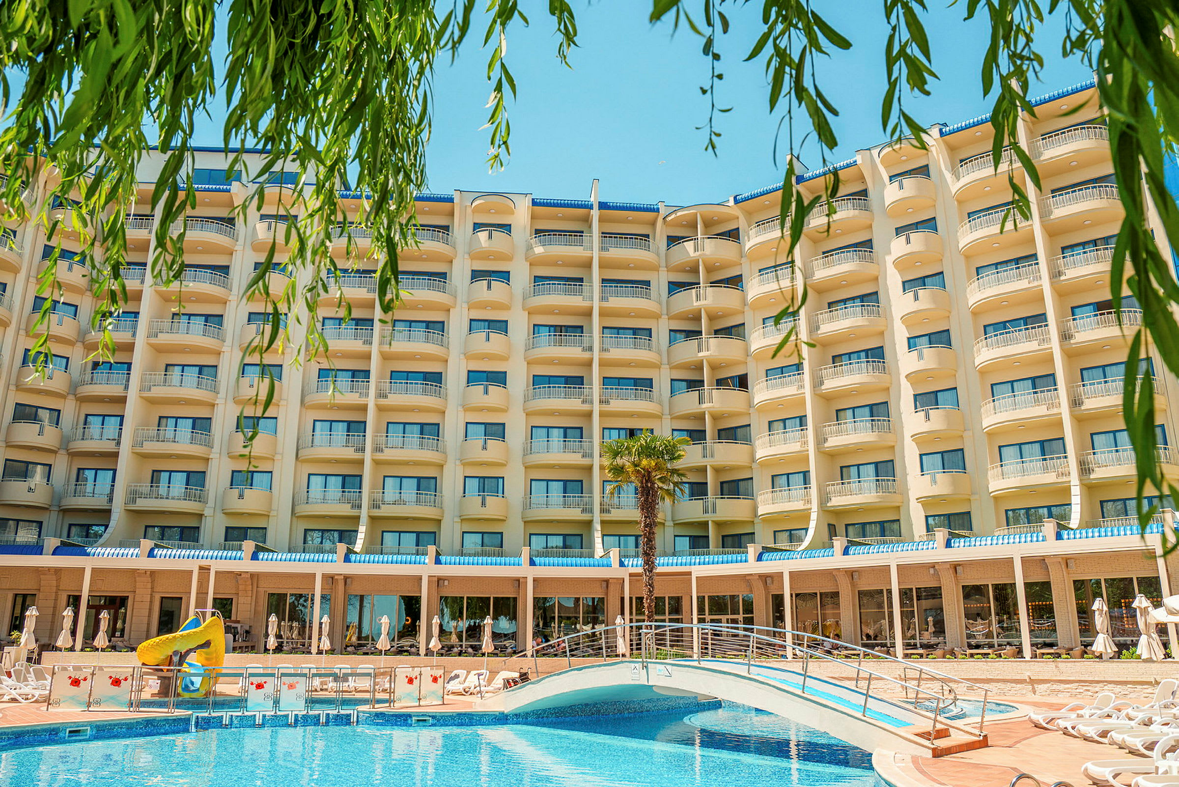 Hotellets fasad med balkonger, en pool med en bro och gröna växter i förgrunden. (AI-genererad text)
