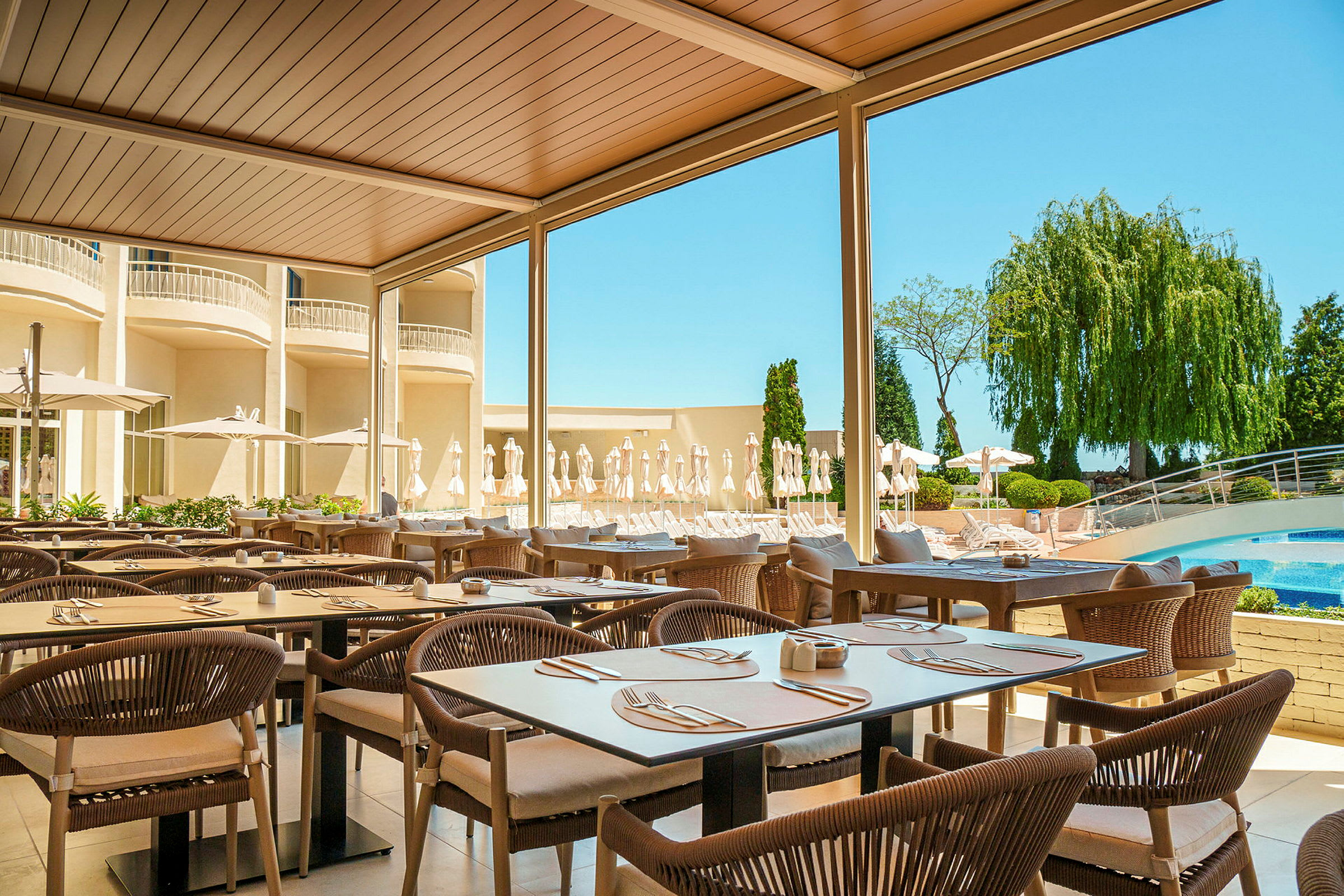 Utomhusrestaurang med bord och stolar under tak, omgiven av grönt och en pool i bakgrunden. (AI-genererad text)