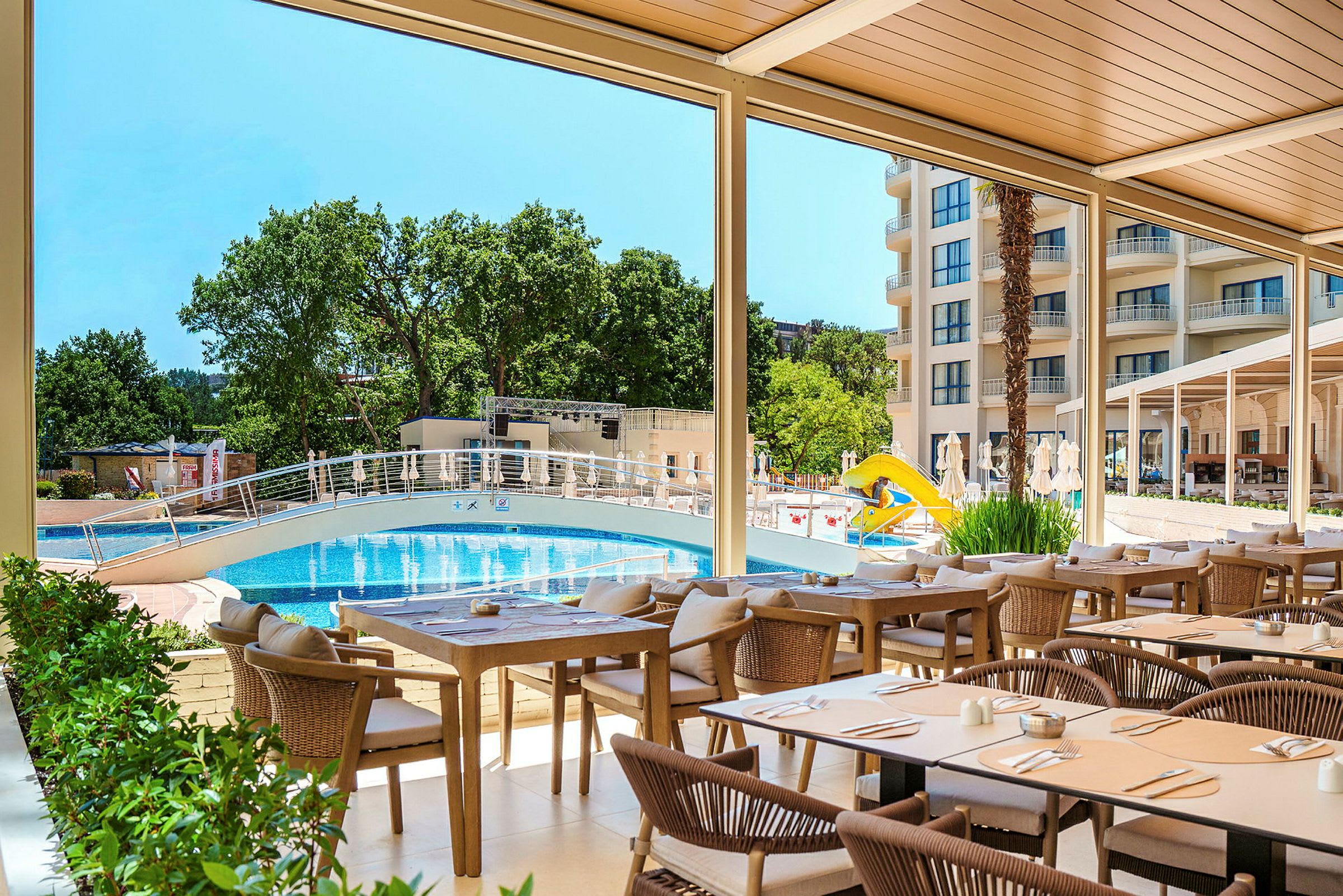 Ett utomhusområde med bord och stolar under ett tak, utsikt över en pool med en bro och en gul rutschbana. Bakom poolen syns ett flervåningshus samt gröna träd. (AI-genererad text)