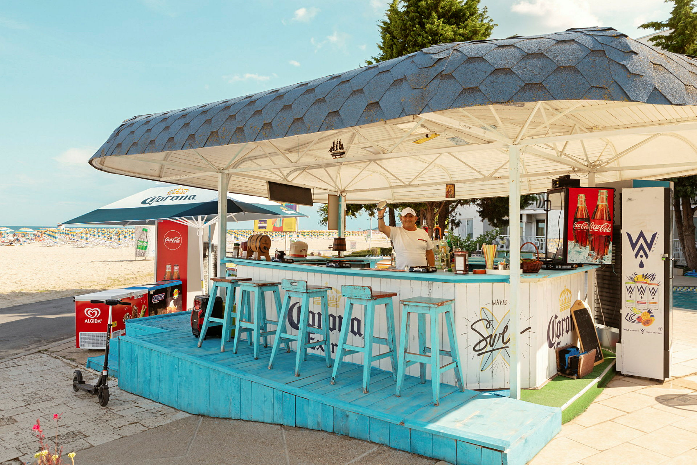 En strandbar med blåa stolar och tak, belägen nära sandstranden. (AI-genererad text)