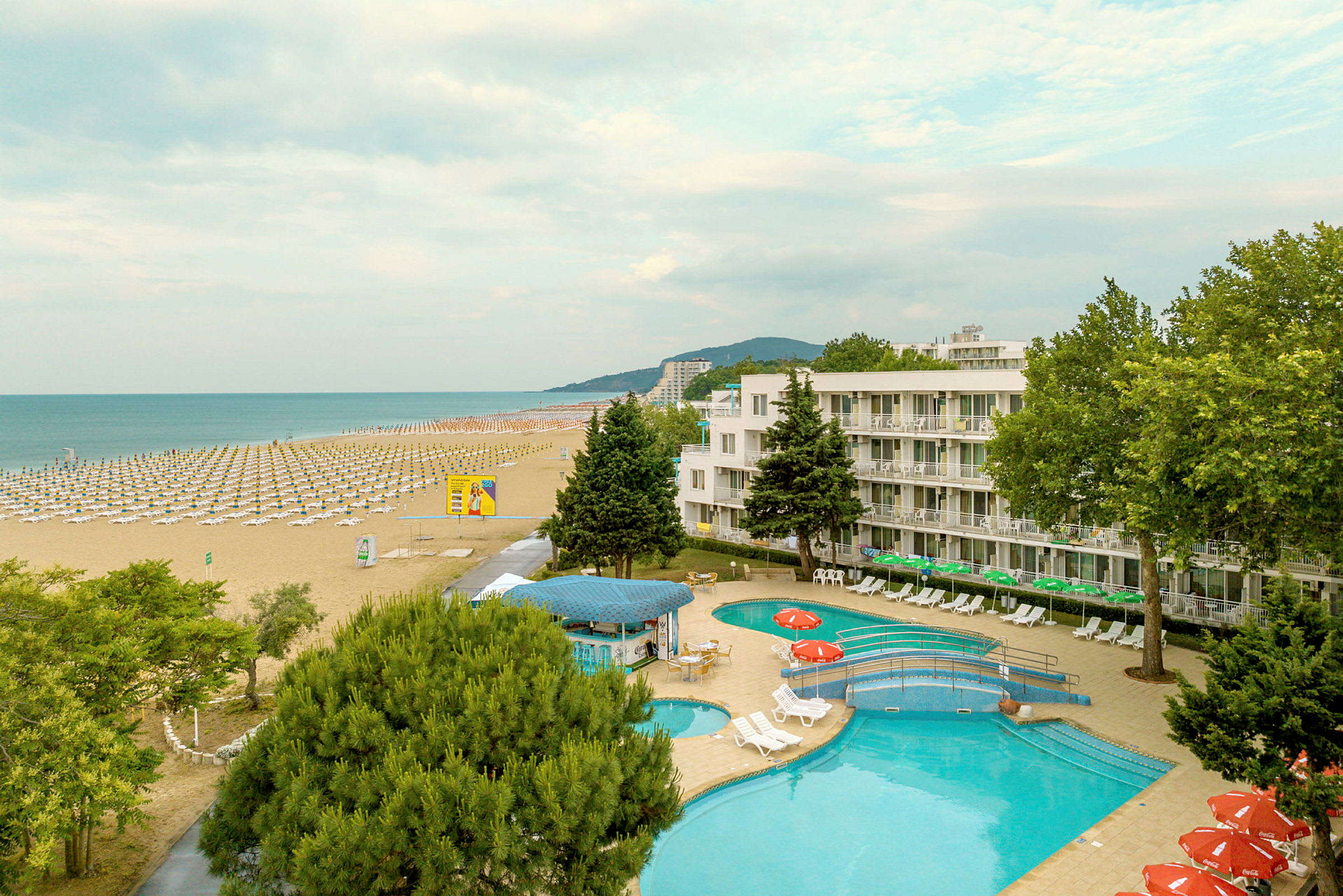 En utsikt över en strand med många solstolar och parasoller, samt ett hotell och en pool i förgrunden. (AI-genererad text)
