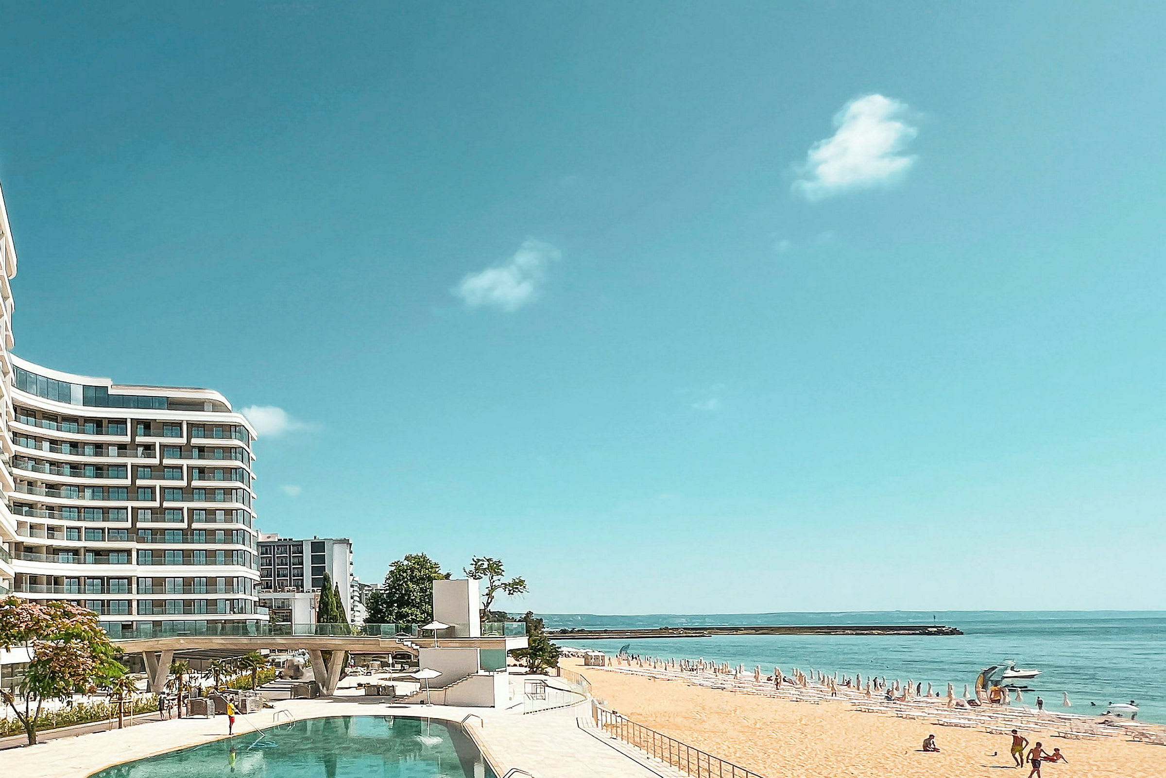 Hotellbyggnad vid en pool och en sandstrand med parasoller vid havet. (AI-genererad text)