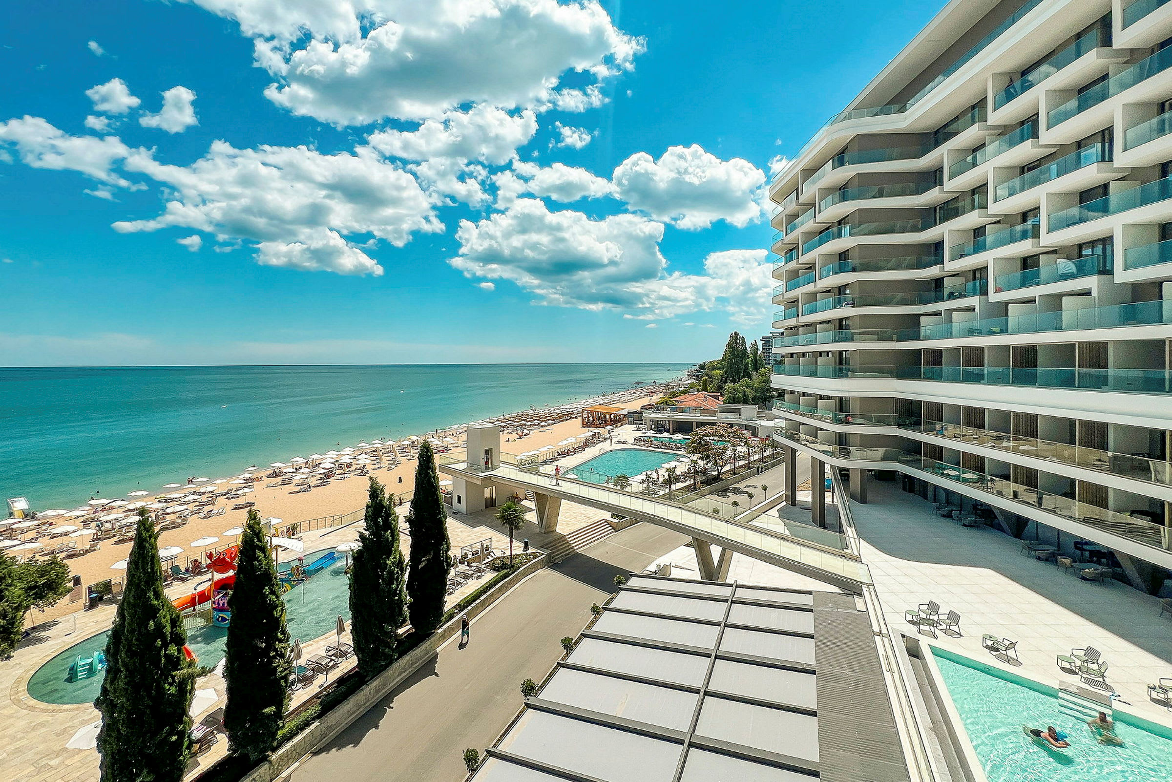 En strand med solstolar och parasoller nära ett hotell med moderna byggnader och poolområden. (AI-genererad text)