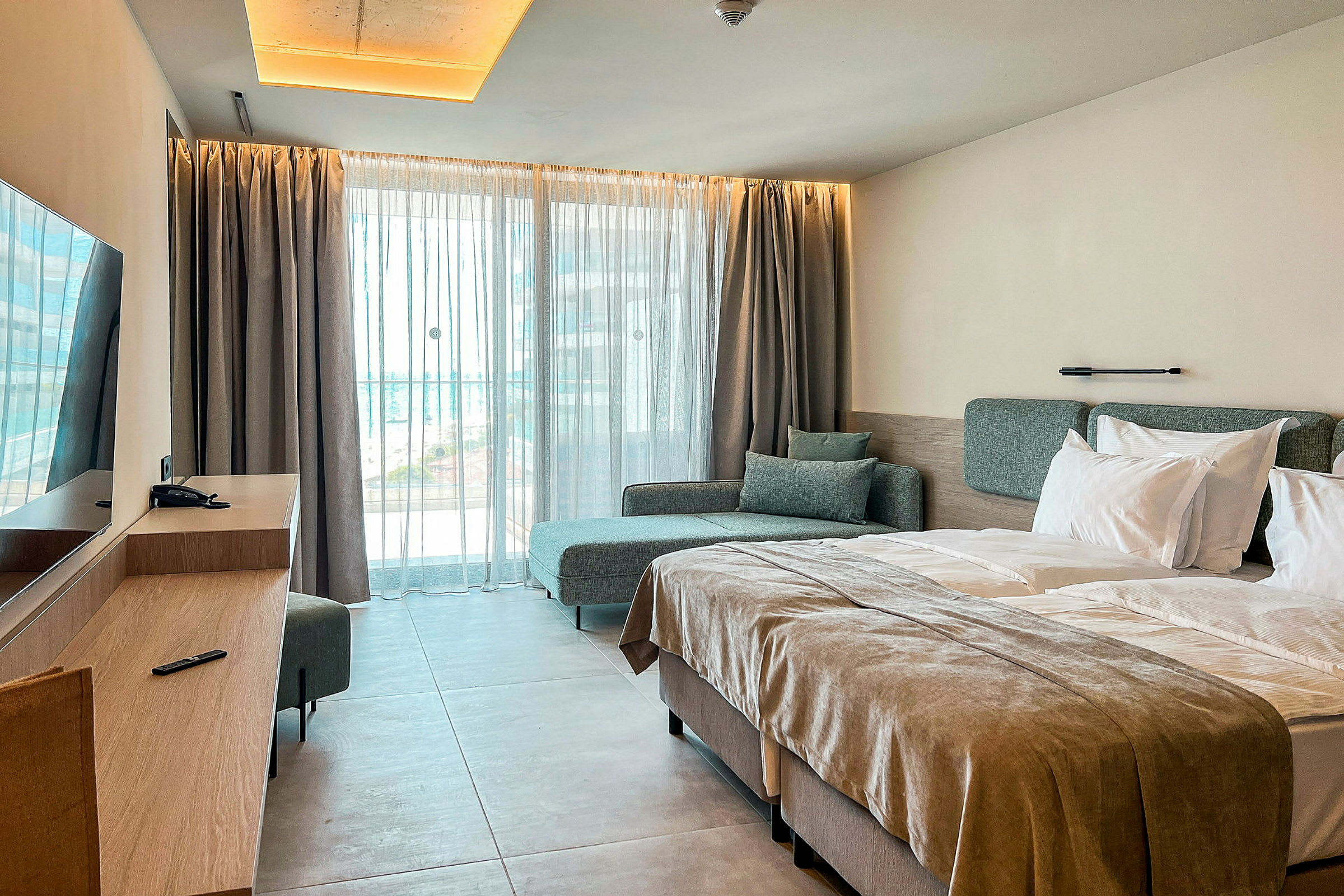 Ett hotellrum med en dubbelsäng, en soffa och ett skrivbord framför ett fönster med ljusa gardiner. (AI-genererad text)