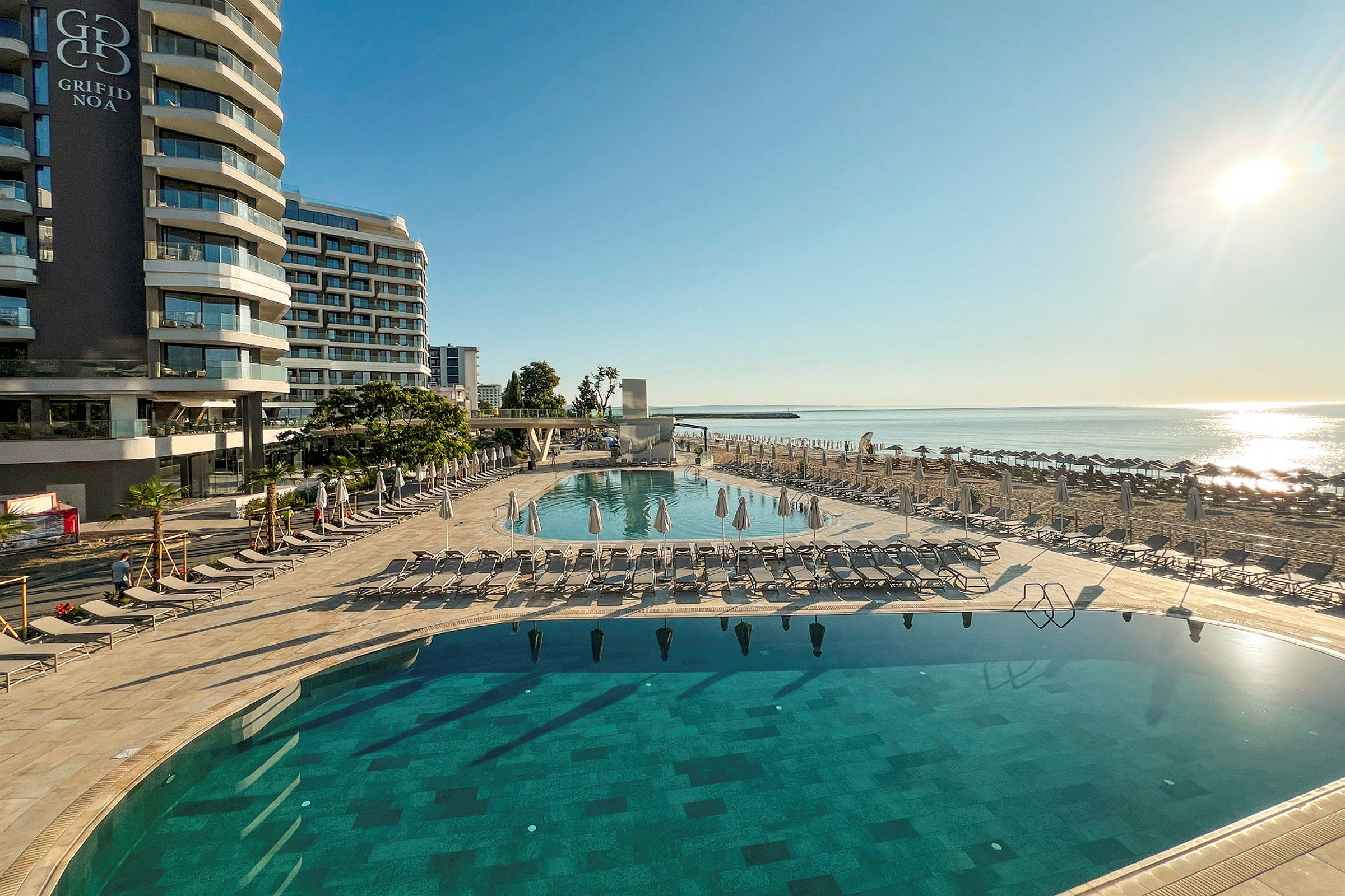 Hotellbyggnad vid strandpromenad med pool och solstolar. (AI-genererad text)