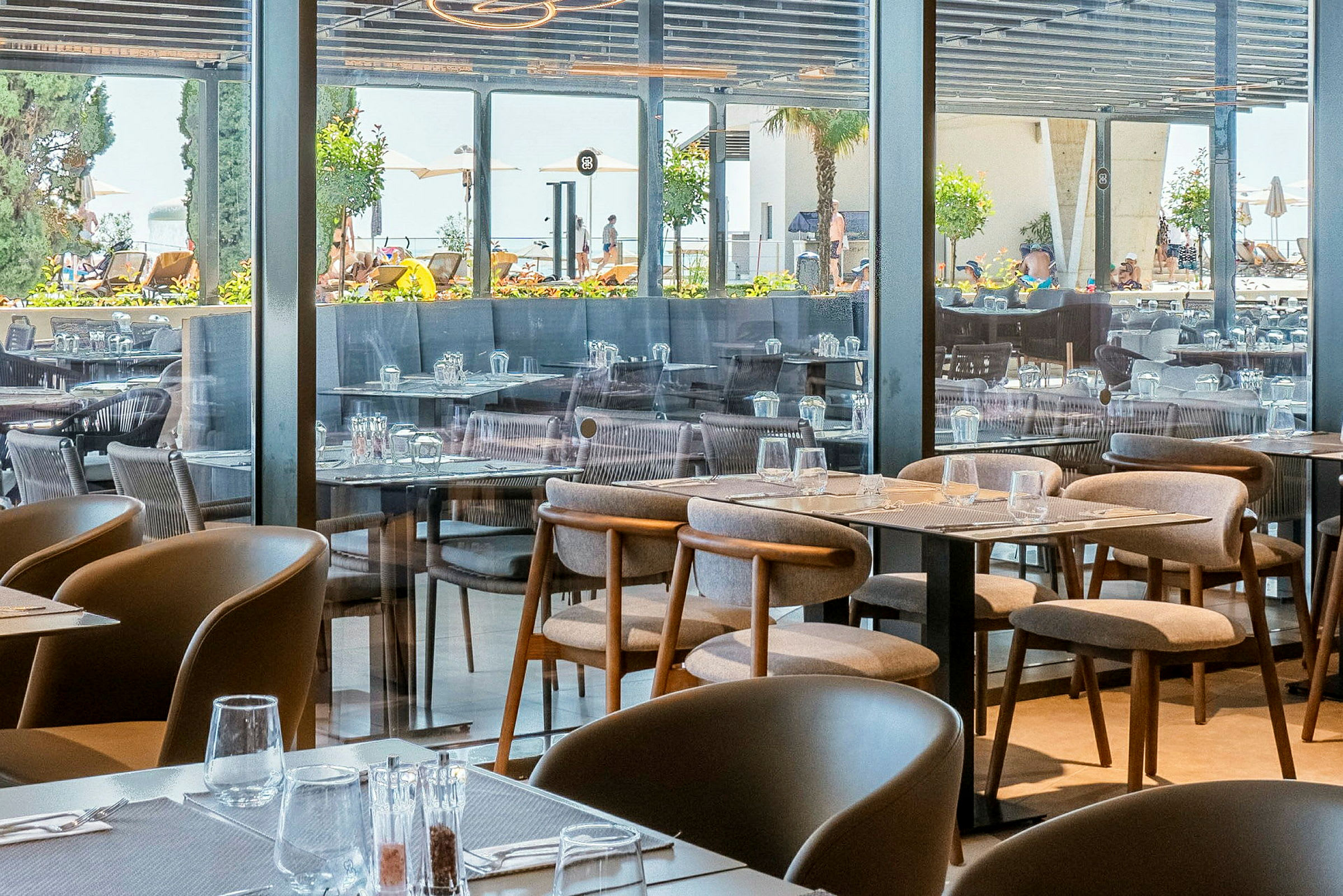 Bild av en restaurang med uppställda bord och stolar, med utsikt mot utomhusområdet genom glasväggar. (AI-genererad text)