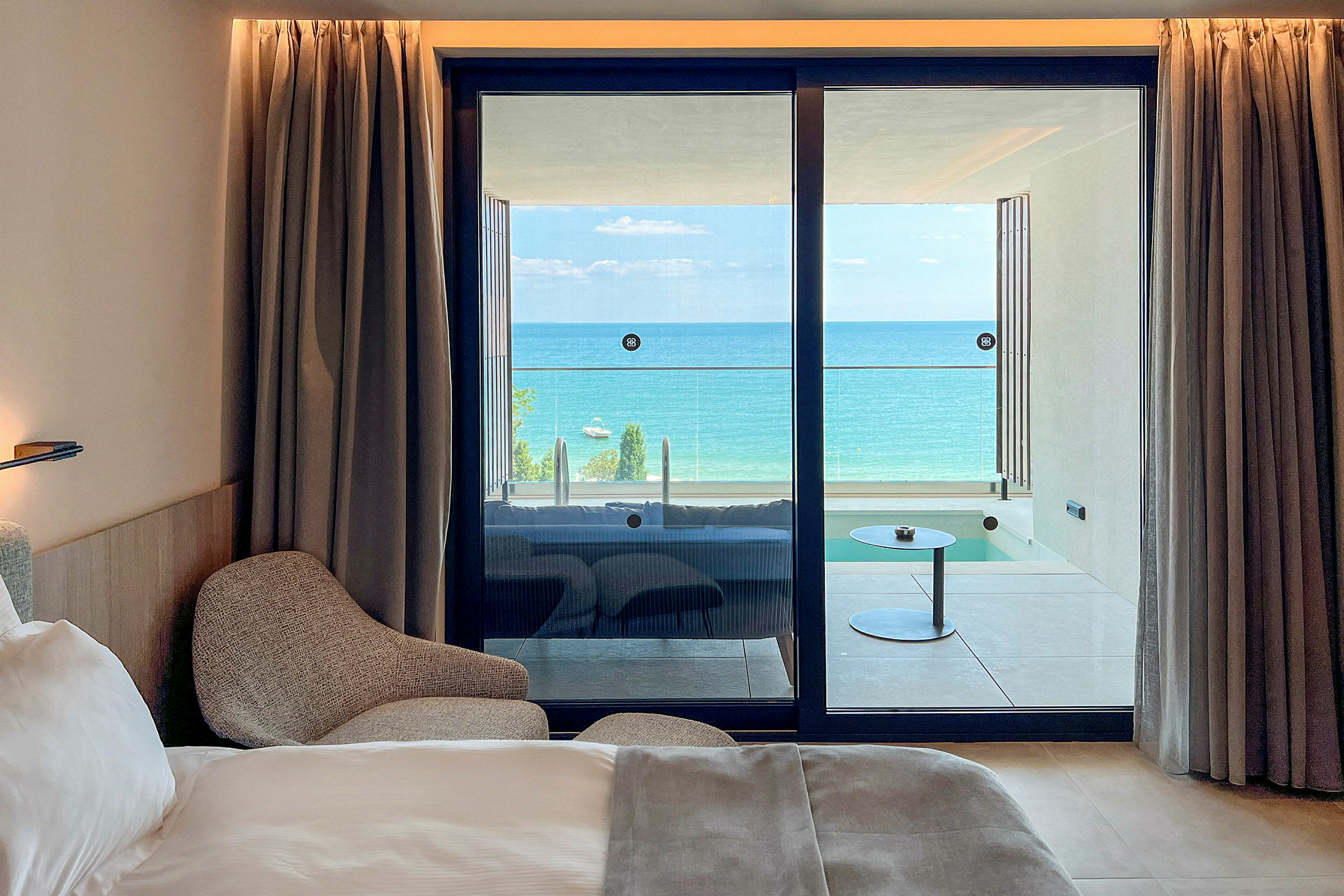 Ett hotellrum med en säng och fåtölj, samt utsikt över en balkong med havet i bakgrunden. (AI-genererad text)