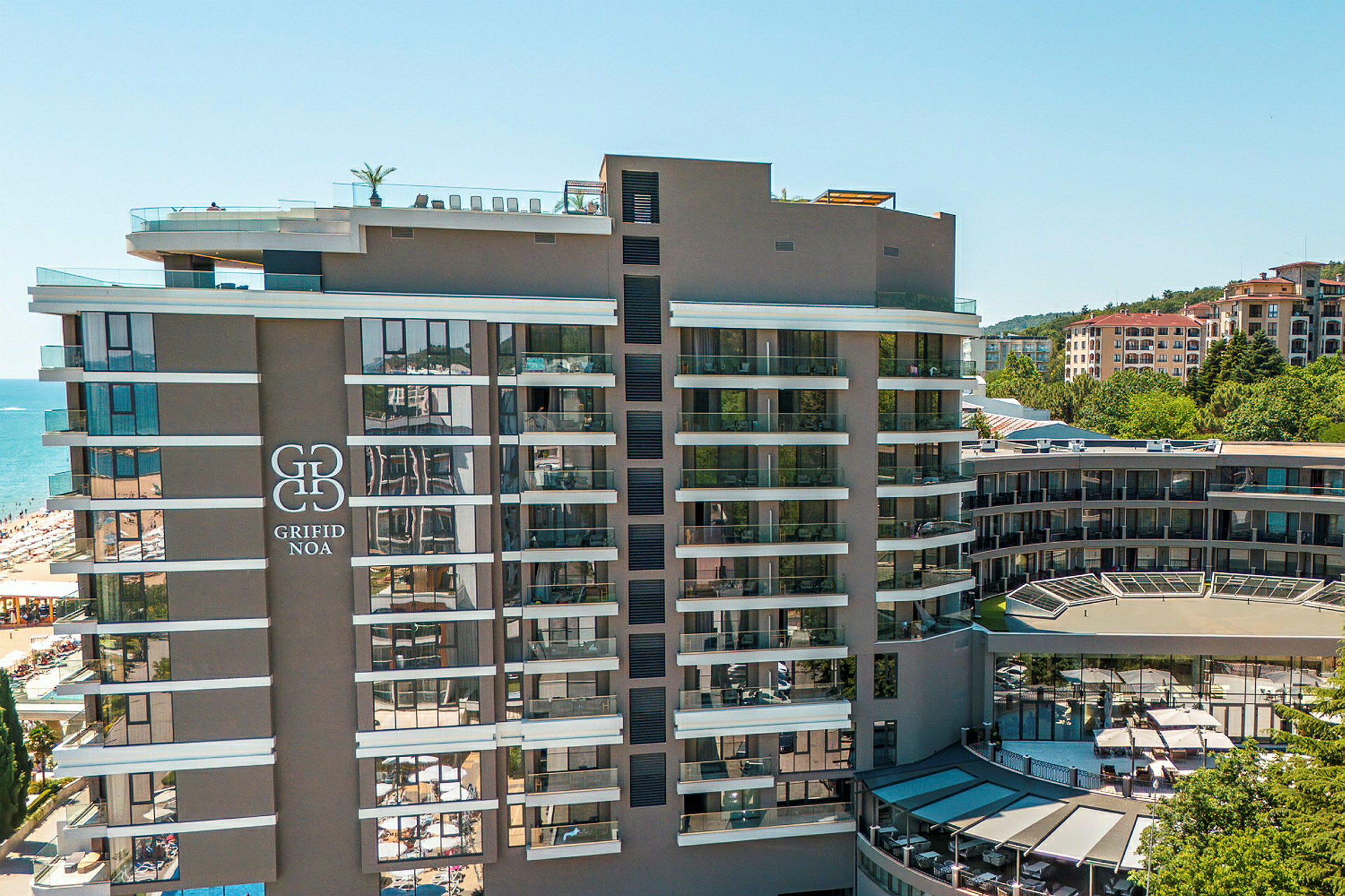 Modernt hotell med flera våningar, stor logotyp på fasaden, beläget nära en strand och omgivande byggnader. (AI-genererad text)