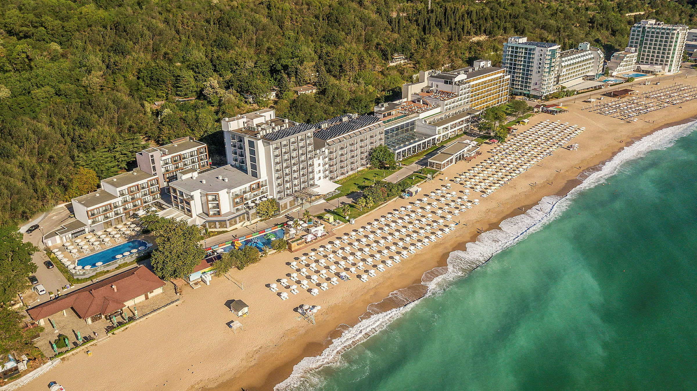 Vy över hotell vid kusten, med strand och parasoller samt grönska i bakgrunden. (AI-genererad text)