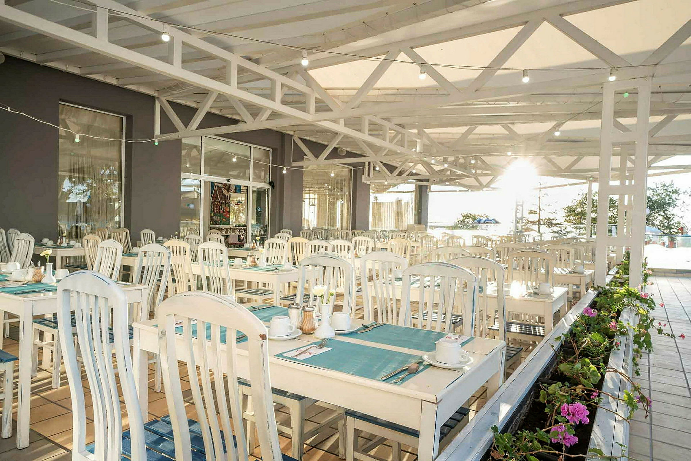 En utomhusrestaurang med bord och stolar i ljus trästil, dukade med tallrikar och koppar, samt planteringar i förgrunden. (AI-genererad text)