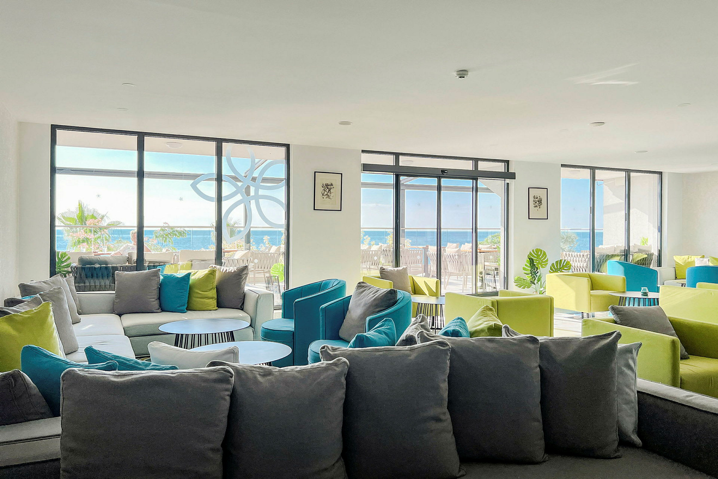 En lounge med soffor och färgglada stolar, och fönster som vetter mot en terrass och havet. (AI-genererad text)