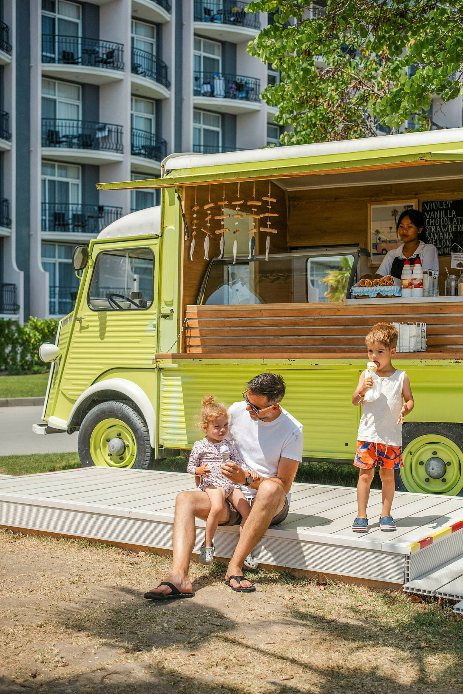 En grön foodtrailer med servering framför en byggnad med balkonger. Två barn och en vuxen sitter på en ramp framför trailern. (AI-genererad text)