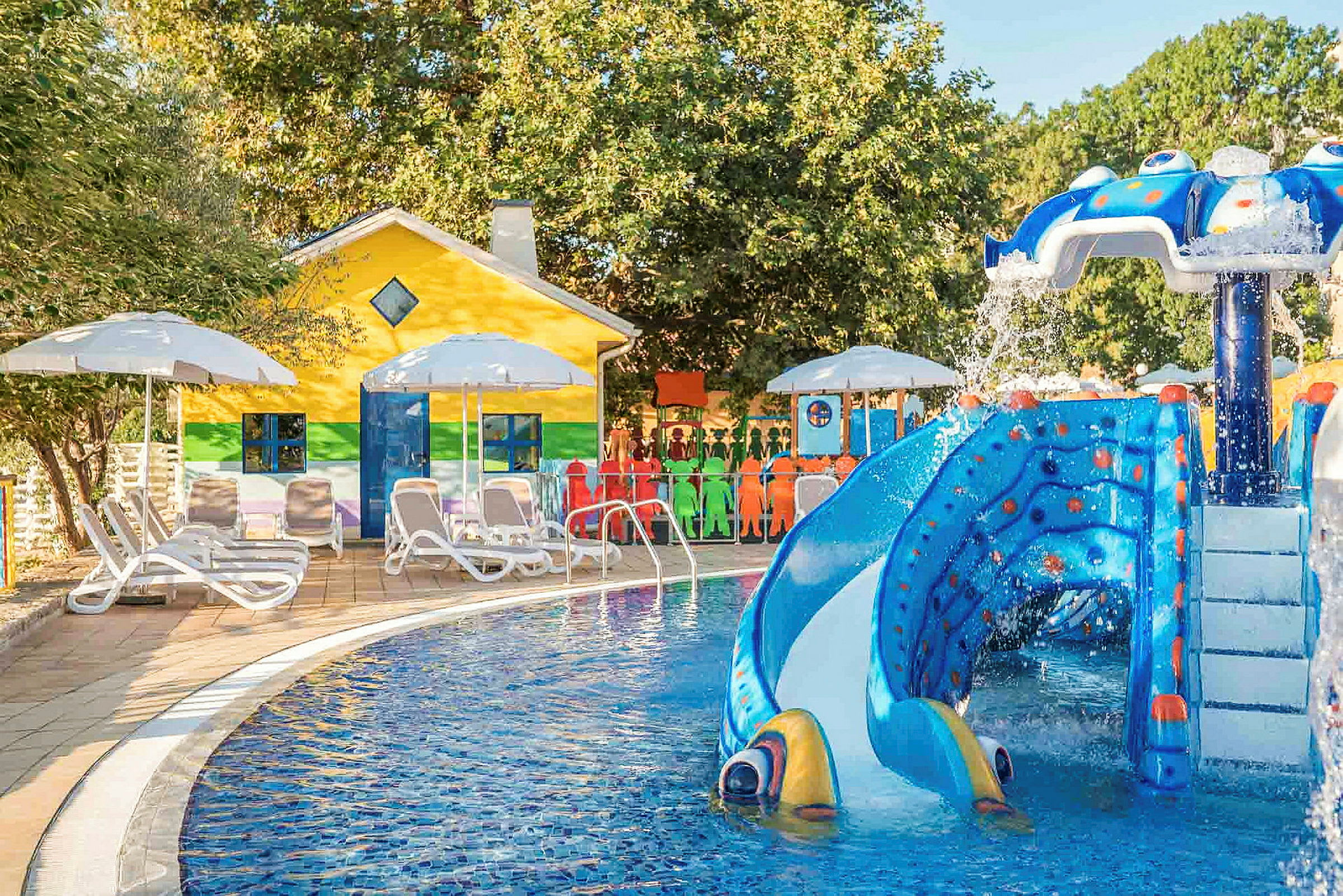 En färgglad byggnad vid sidan av en pool med vattenrutschbana och parasoller. (AI-genererad text)