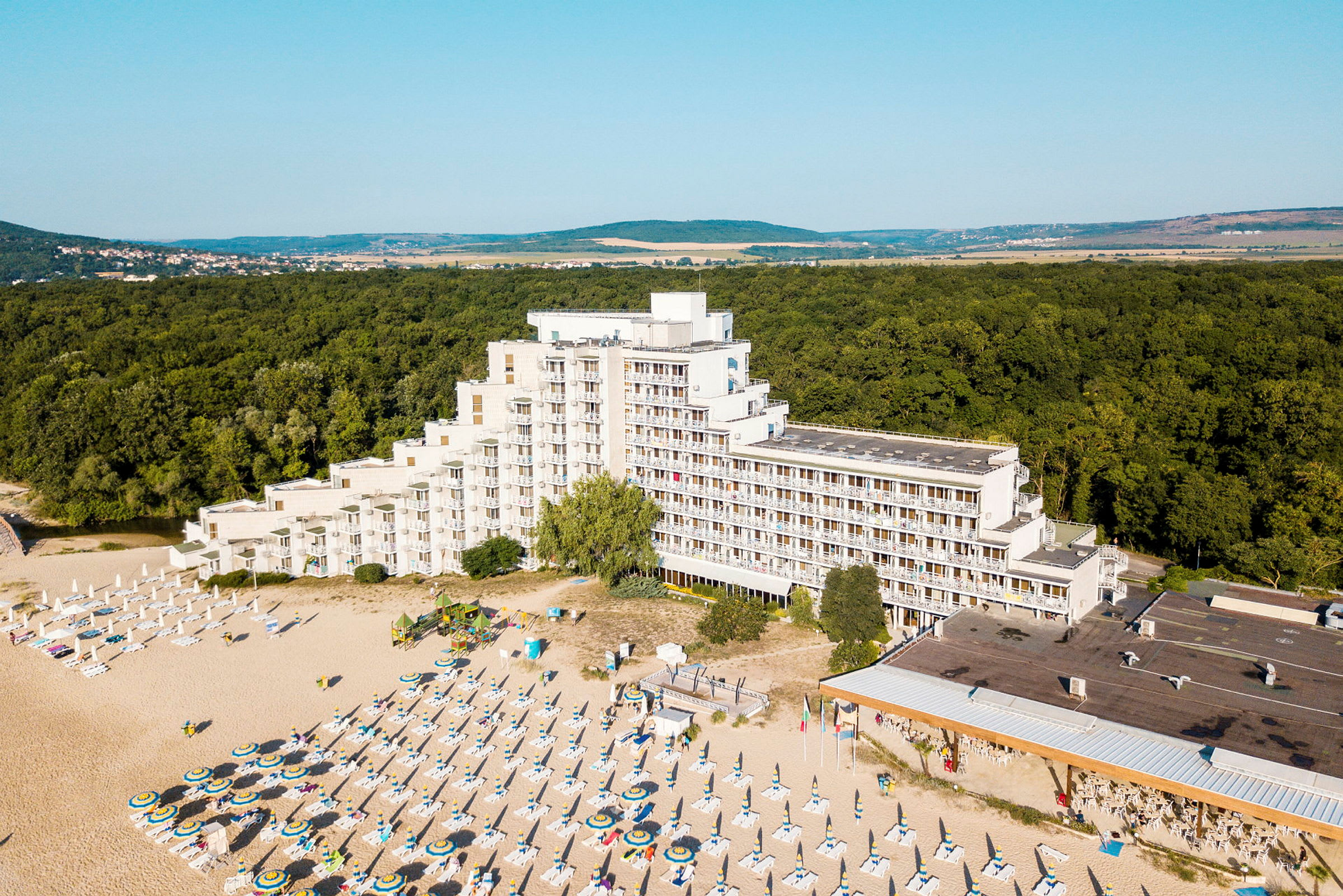 Bild av ett hotell vid en strand med solstolar och parasoller, omgiven av skog. (AI-genererad text)