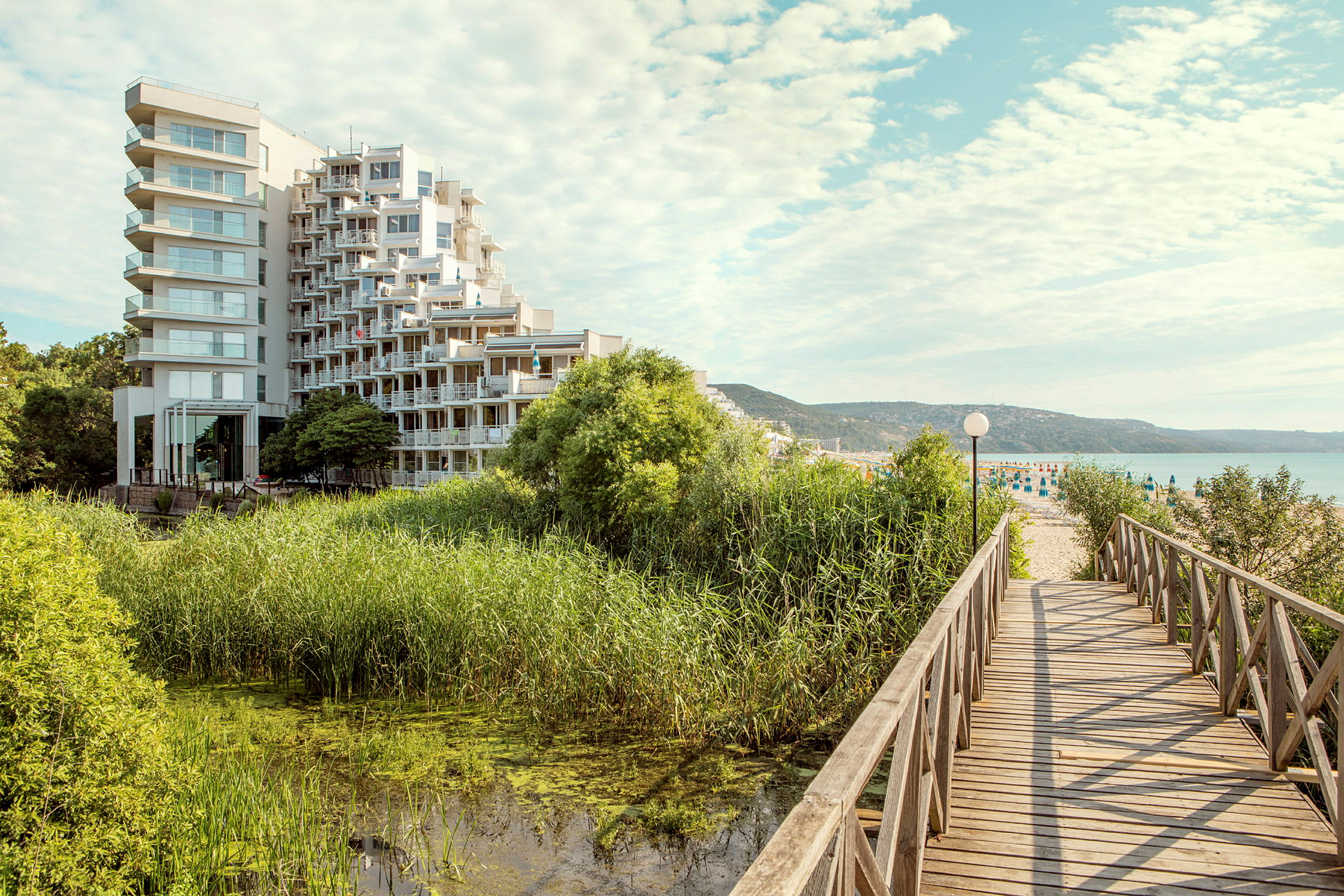 En modern hotellbyggnad ligger nära ett område med grönska och vatten, och en träbro leder mot en sandstrand. (AI-genererad text)