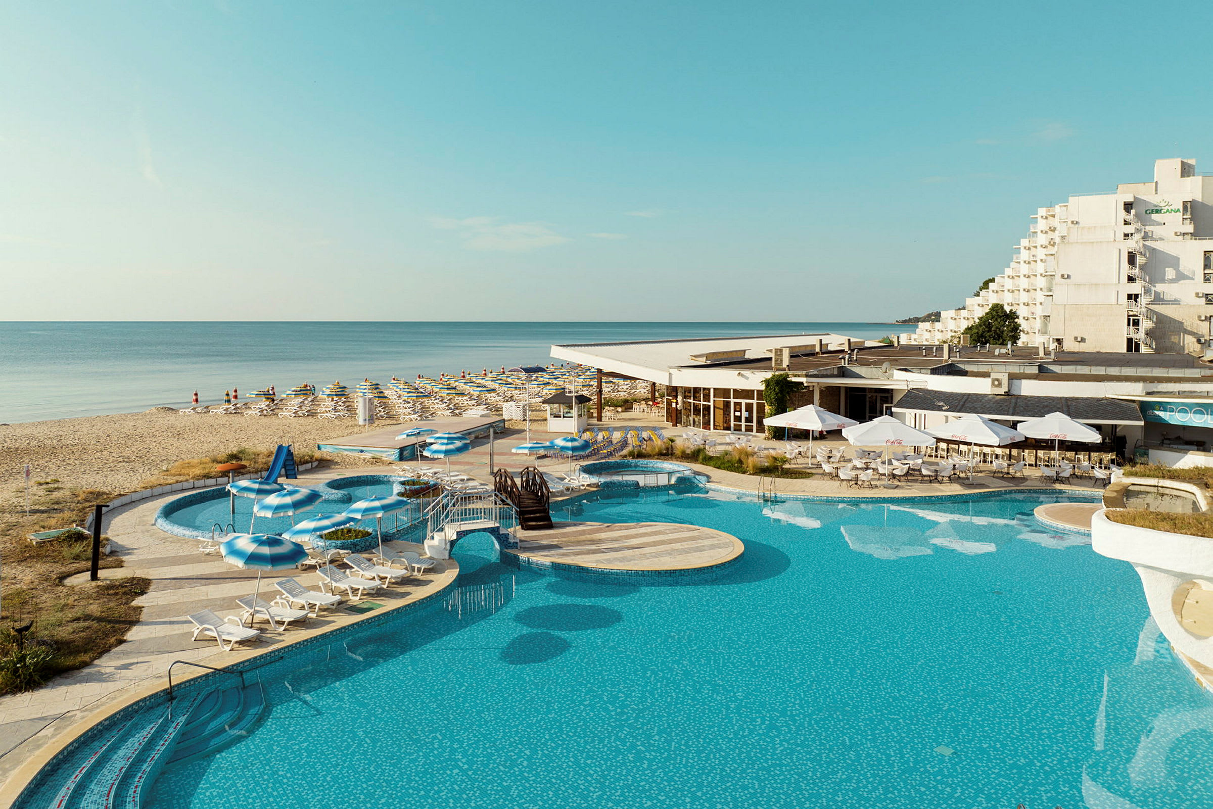 En bild av en pool, solstolar, och parasoller vid ett hotell nära en sandstrand. (AI-genererad text)